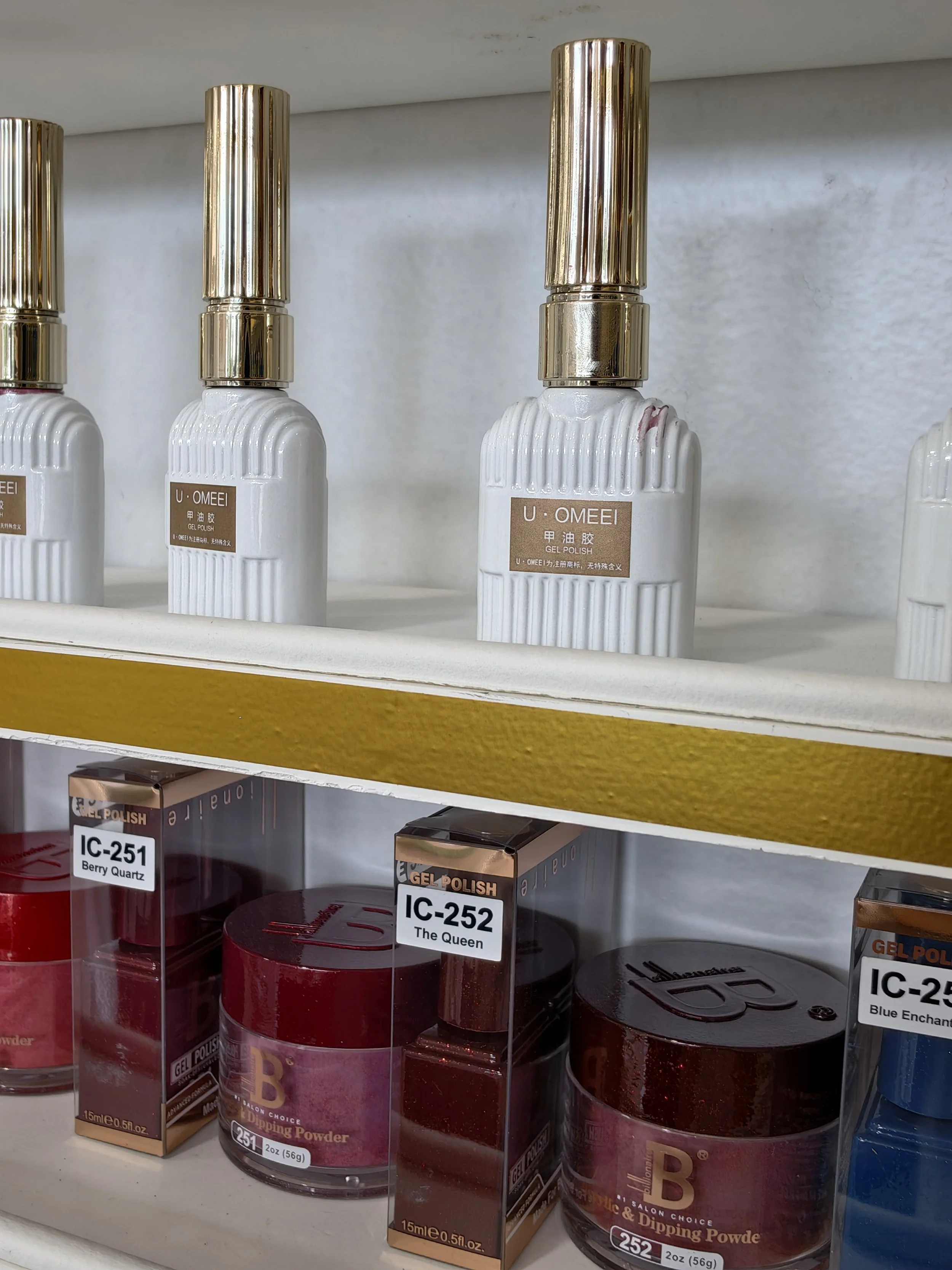 Various bottles of gel nail polish with gold caps and labels, displayed on store shelves. Some bottles are labeled 'U-OMEI' and 'Dipping Powder'.