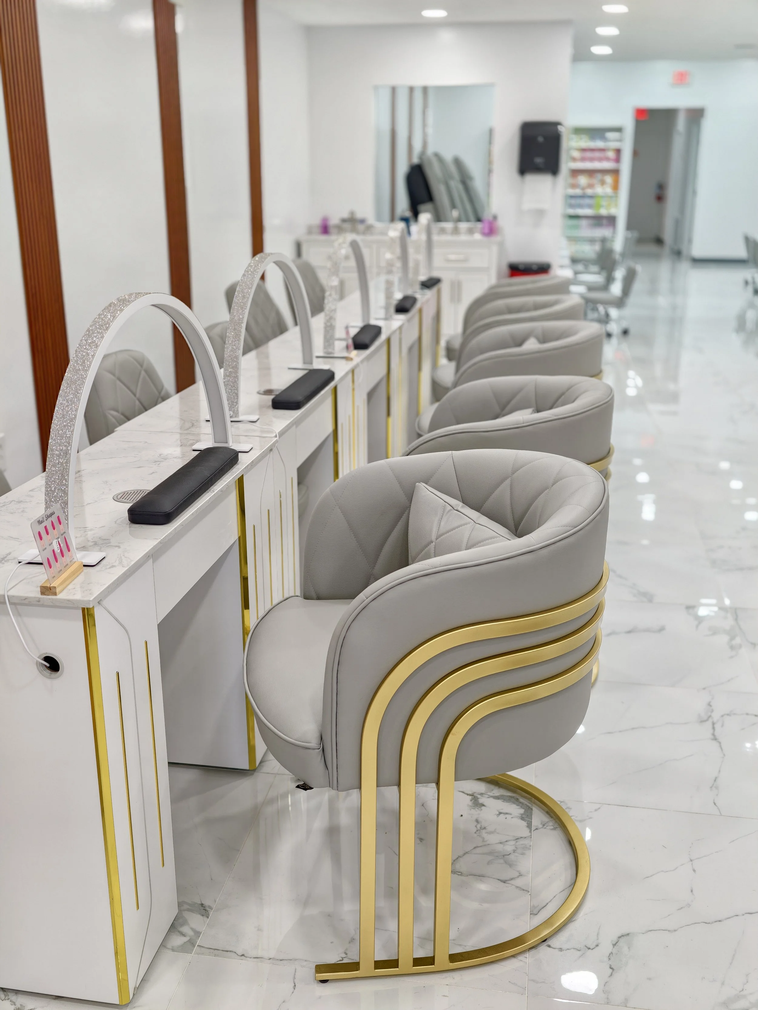 A row of white manicure stations with gold accents, each with a cushioned chair and a nail drying lamp, in a clean salon with a marble floor.
