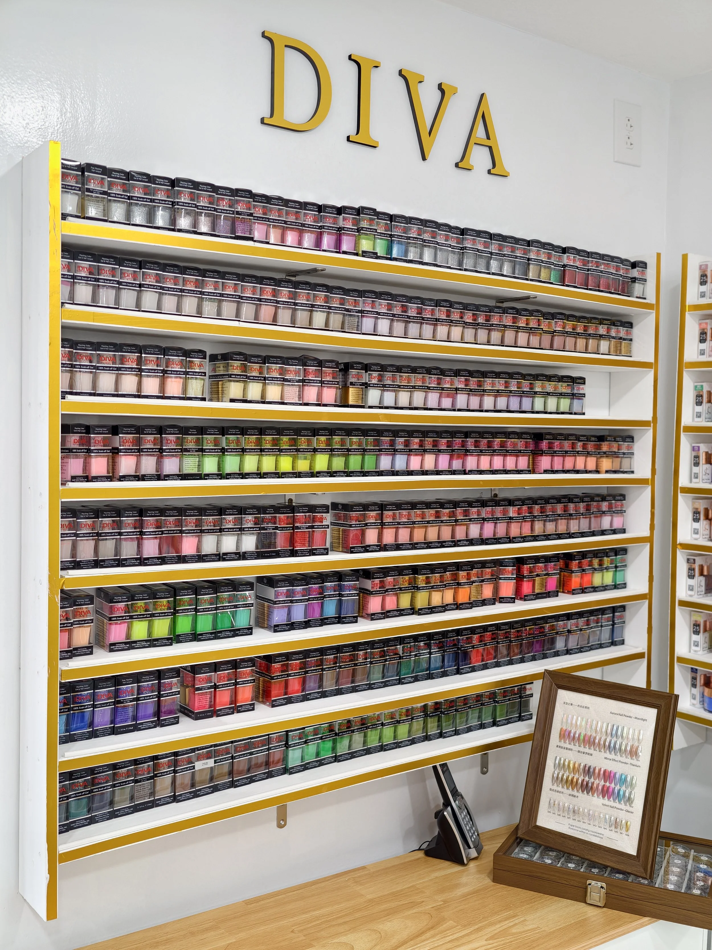 Display of various shades and colors of DIVA nail powders on shelves in a store, with a framed color chart on a wooden table below, and a phone and sample tray nearby.