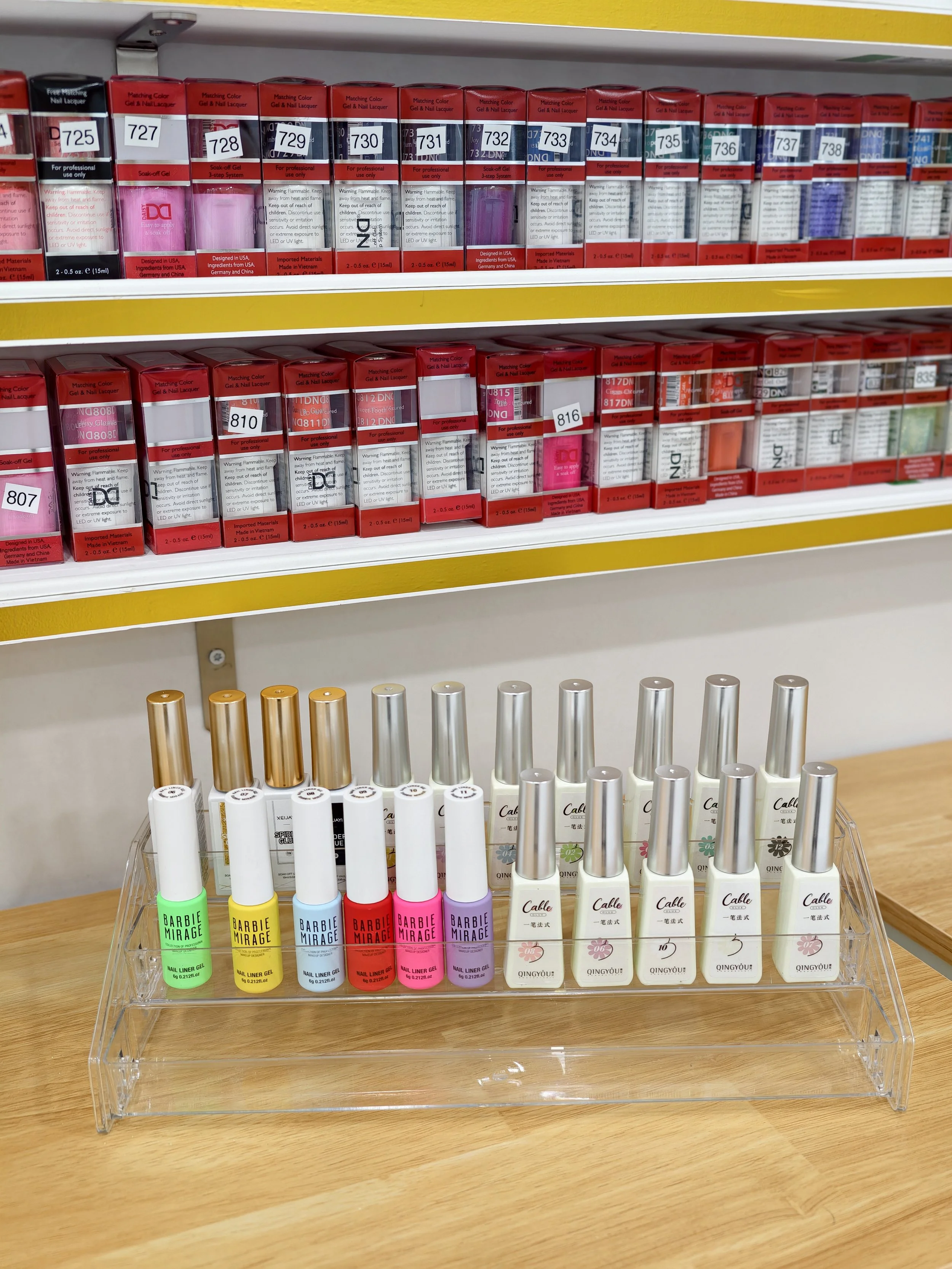 A display stand with colored nail polish and lip gloss bottles on a wooden surface. The stand contains five lip gloss bottles labeled 'QINGYOU' with floral designs, and five nail polish bottles labeled 'Barbie MIRAGE' in bright colors including green