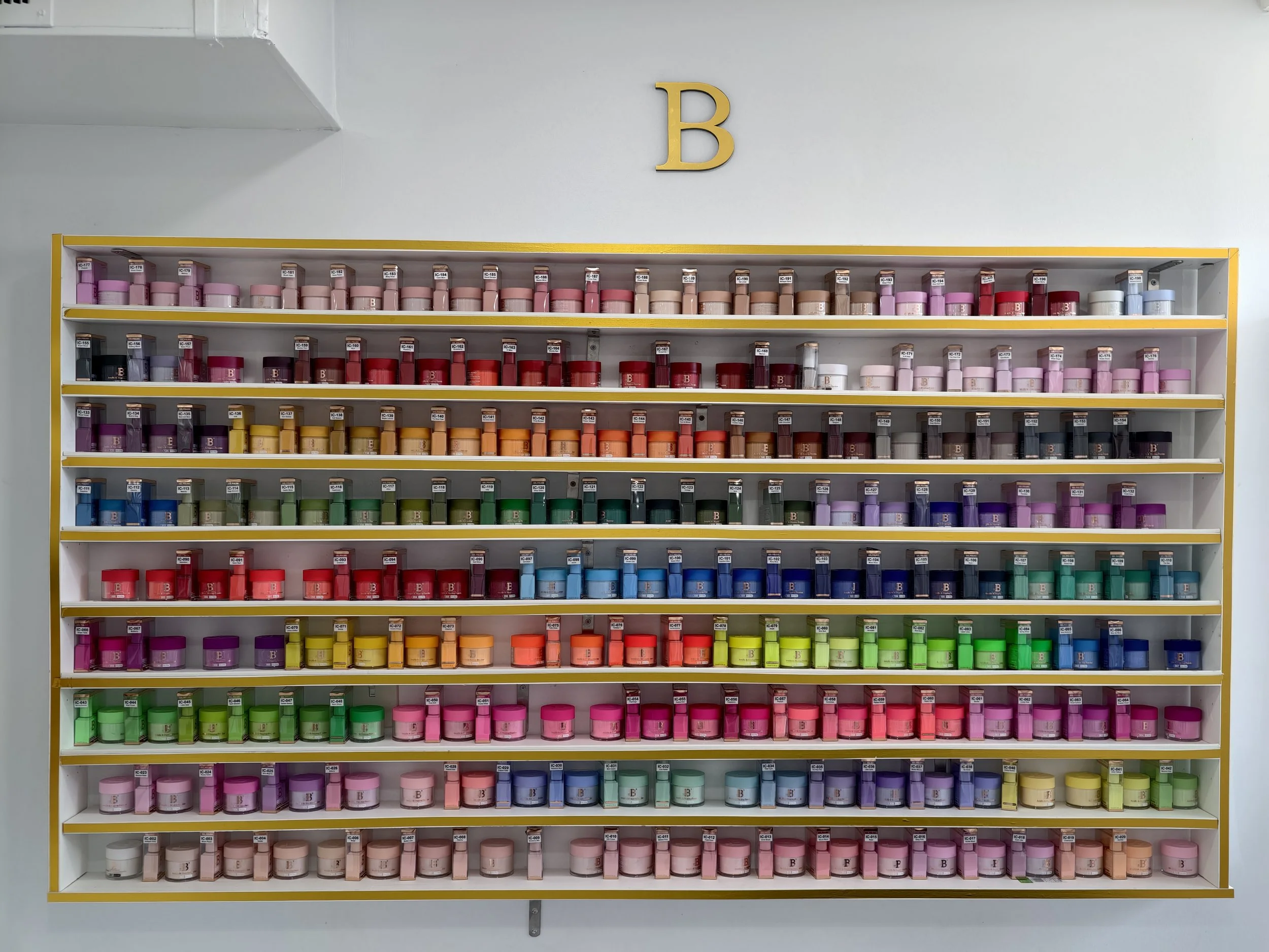 Colorful shelf of paint cans arranged by color in a store, with a large yellow letter 'B' on the wall above.