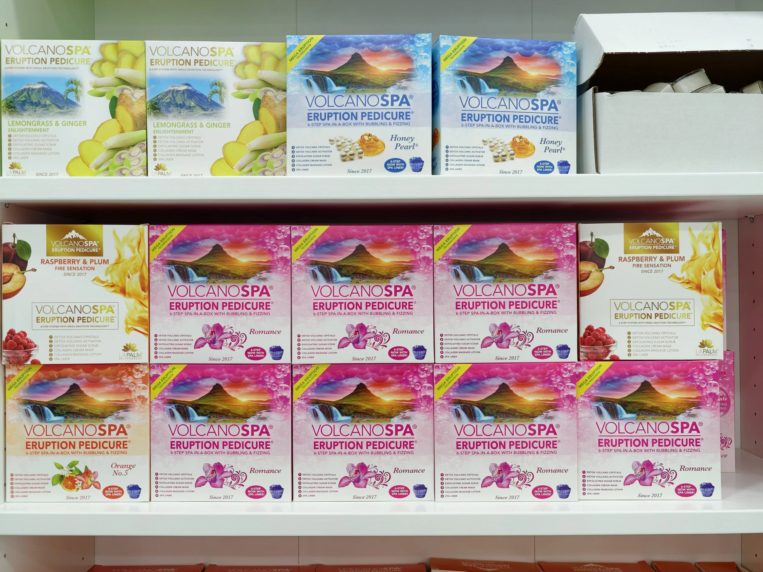 Shelves displaying boxes of Volcano Spa Eruption Pedicure products in various scents, including Lemon Grass & Ginger, Honey Pearl, Raspberry & Plum, and Orange flavors, with colorful mountain and waterfall landscape graphics on the packaging.