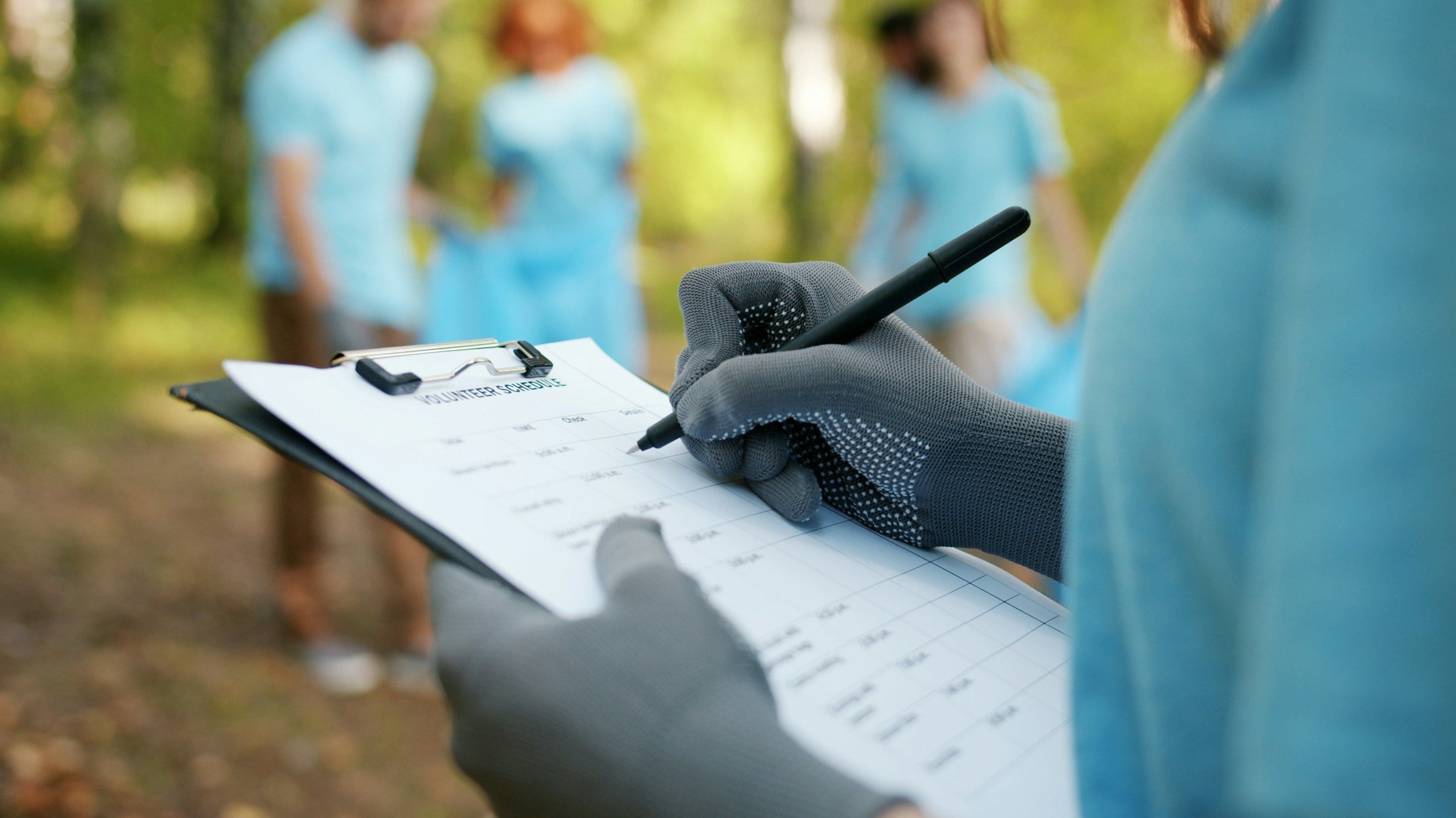 Person in gray gloves filling out a volunteer schedule on a clipboard, in an outdoor park, with three women in light blue shirts in the background.