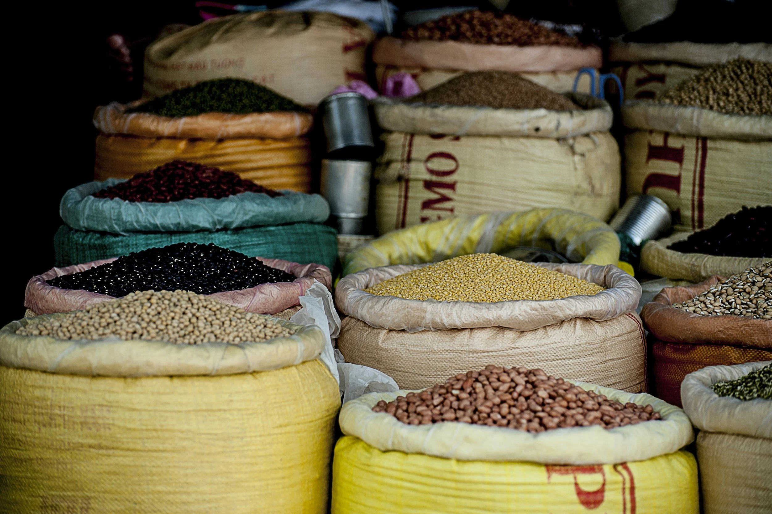 Bags of various grains and legumes displayed at a market, including beans, lentils, and peas in different colored bags.