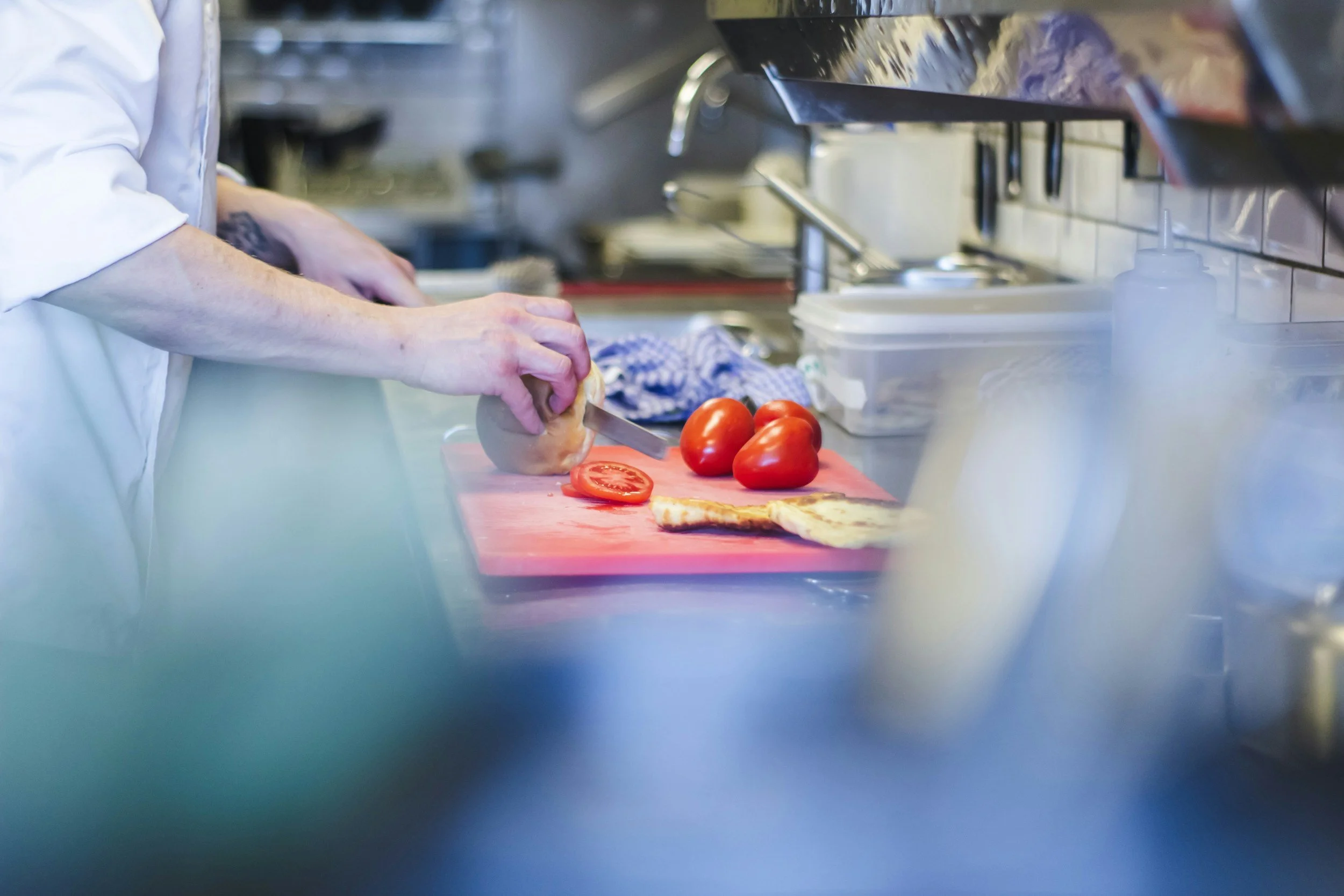A person wearing a white shirt slicing a tomato and a piece of bread on a red cutting board in a kitchen.