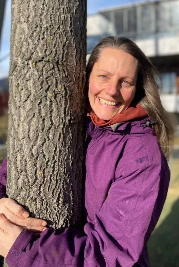 Woman with long hair smiling and hugging a tree outside, wearing a purple jacket and a brown scarf.