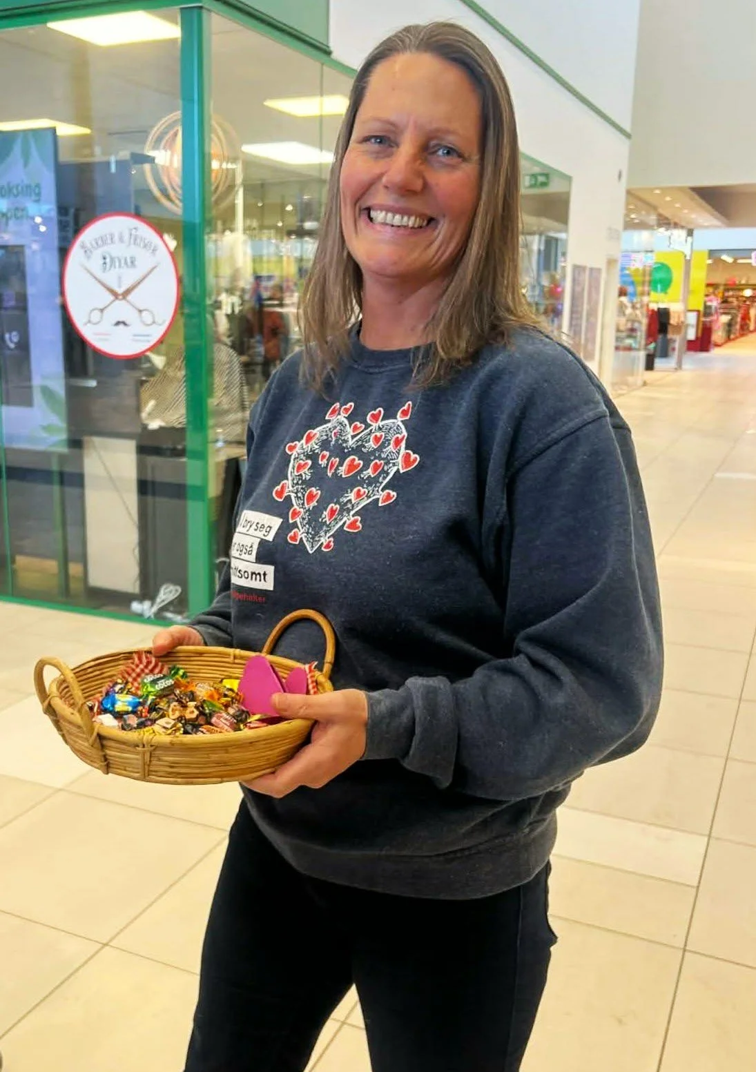 Woman smiling, holding a basket of assorted candies inside a shopping mall.