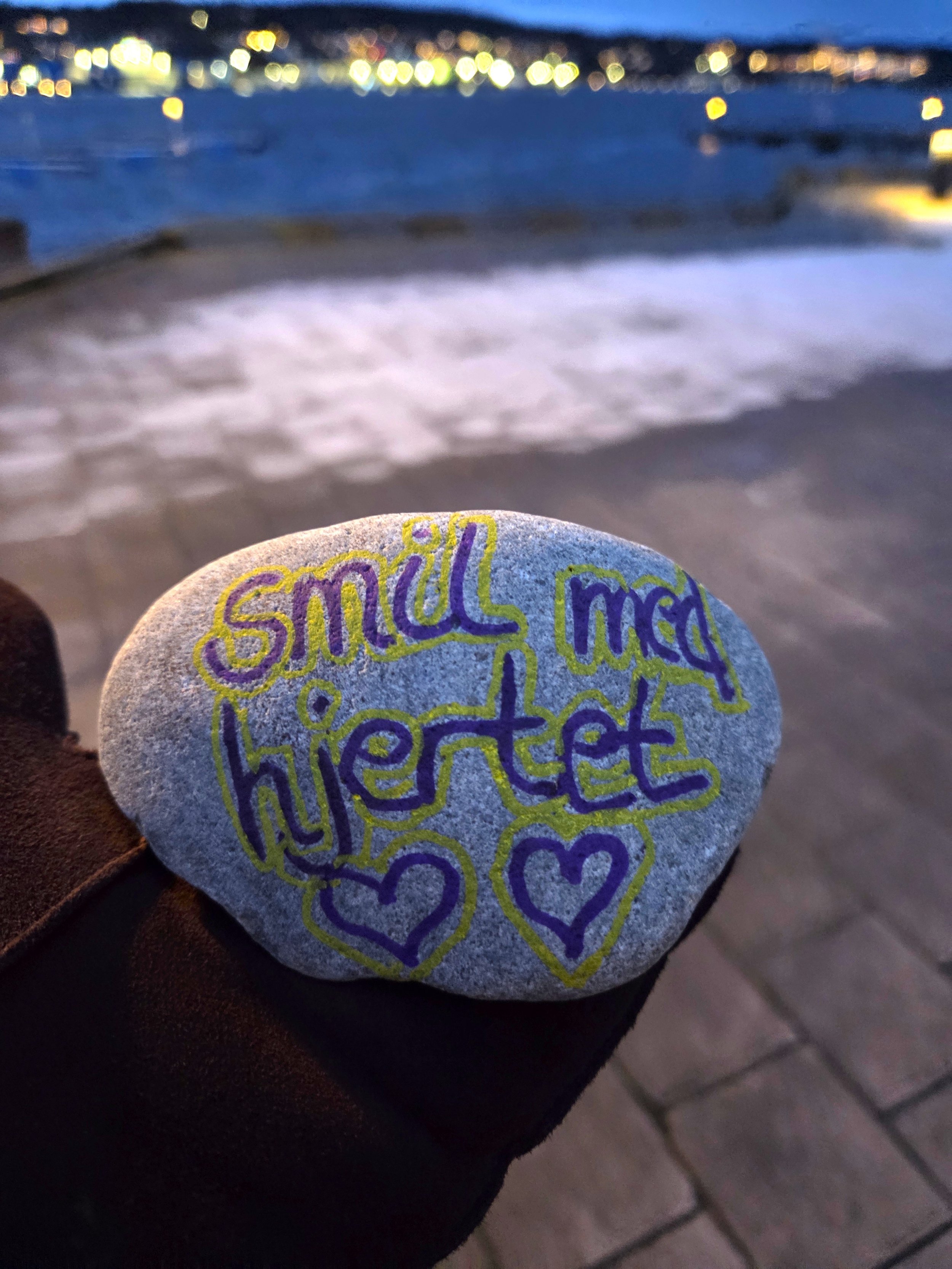A small painted rock with colorful writing reading 'small acts' and heart symbols, held against a beach background during dusk or night.