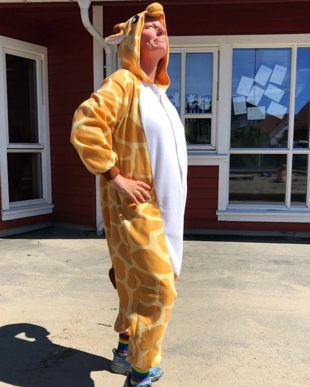 Person standing outdoors in a giraffe costume, looking upward, with a weathered red house and windows behind.