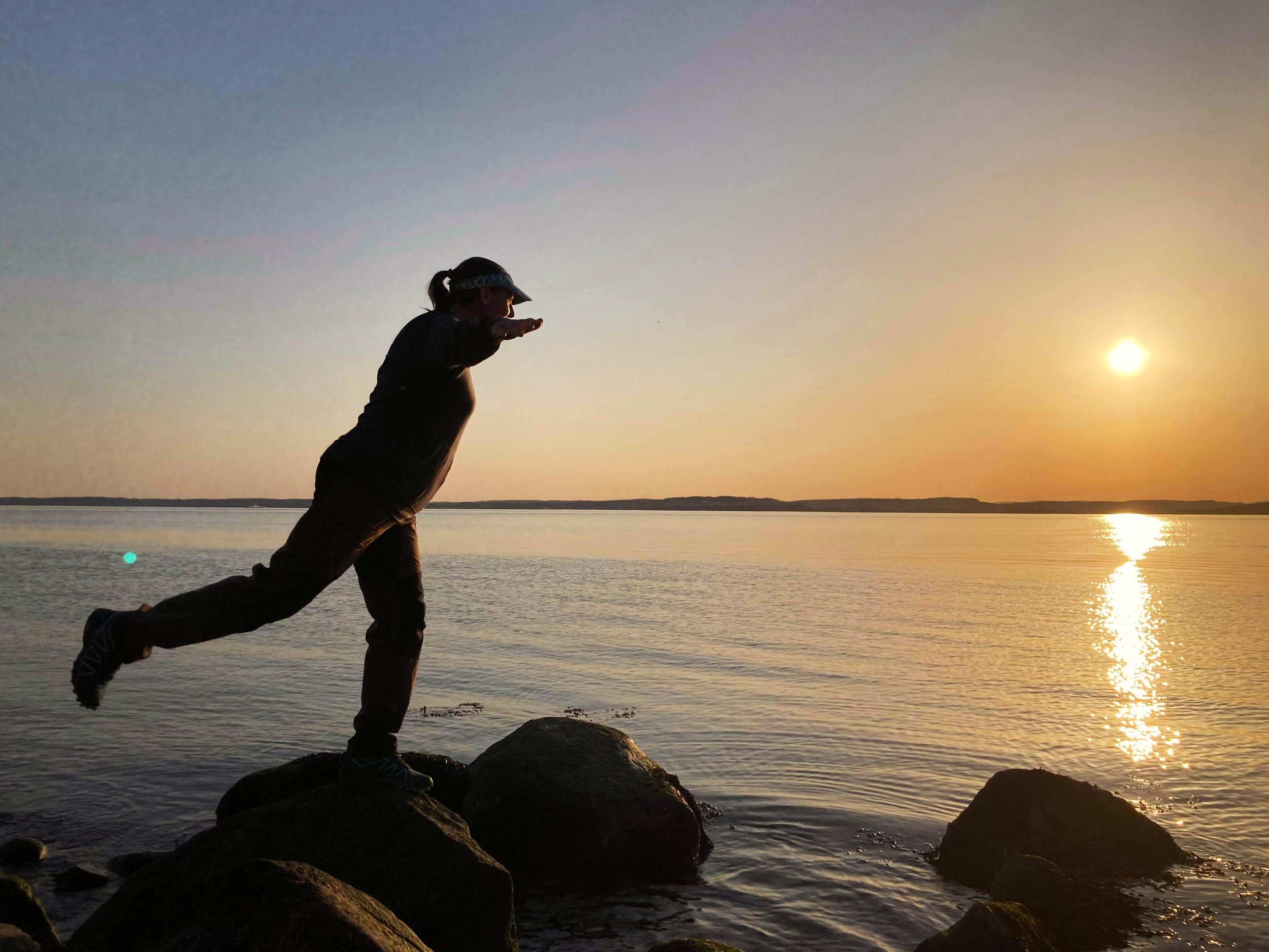 Person running on rocks along a body of water during sunset, with the sky painted in warm colors and the sun reflecting on the water.
