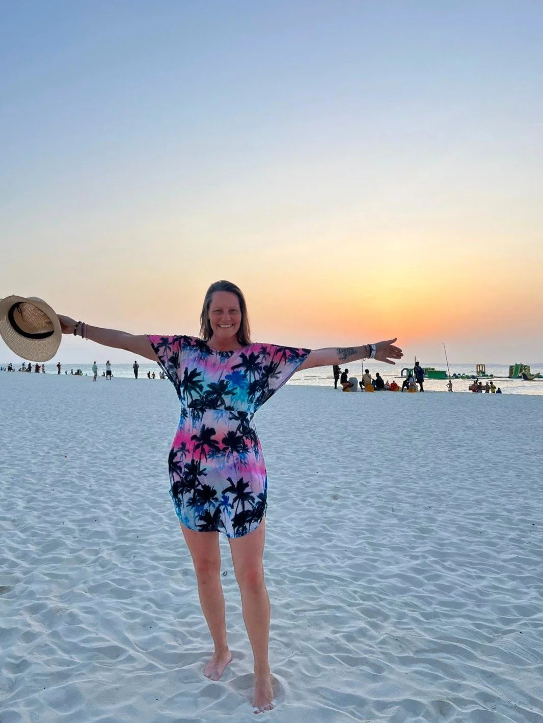 Woman in a colorful tropical dress balances with arms outstretched on a sandy beach during sunset, holding a straw hat in her left hand.