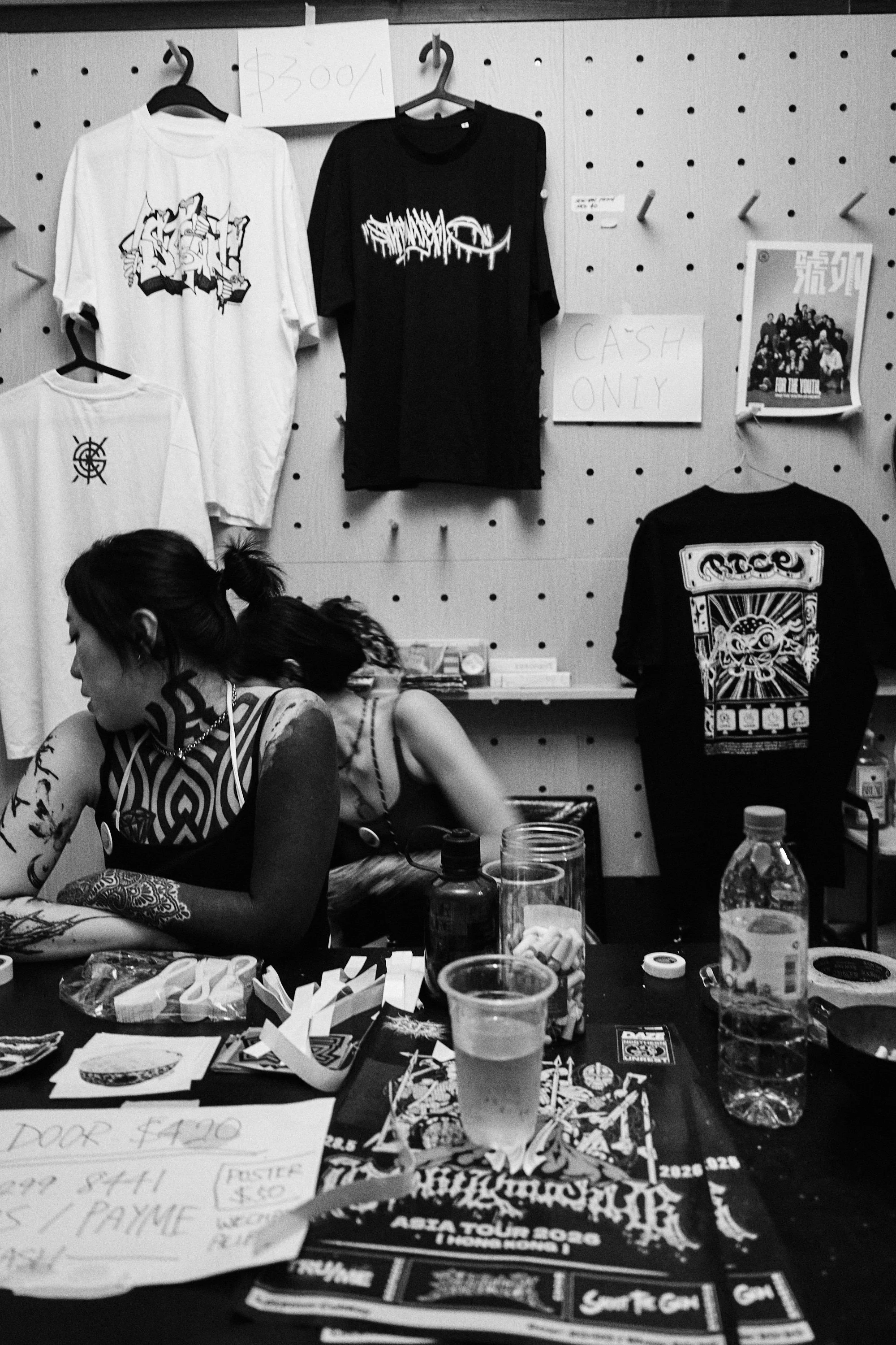 Black-and-white photo of a wall displaying graphic T-shirts on hangers, with handwritten price signs and a magazine, along with a table cluttered with papers, cards, and drink containers.