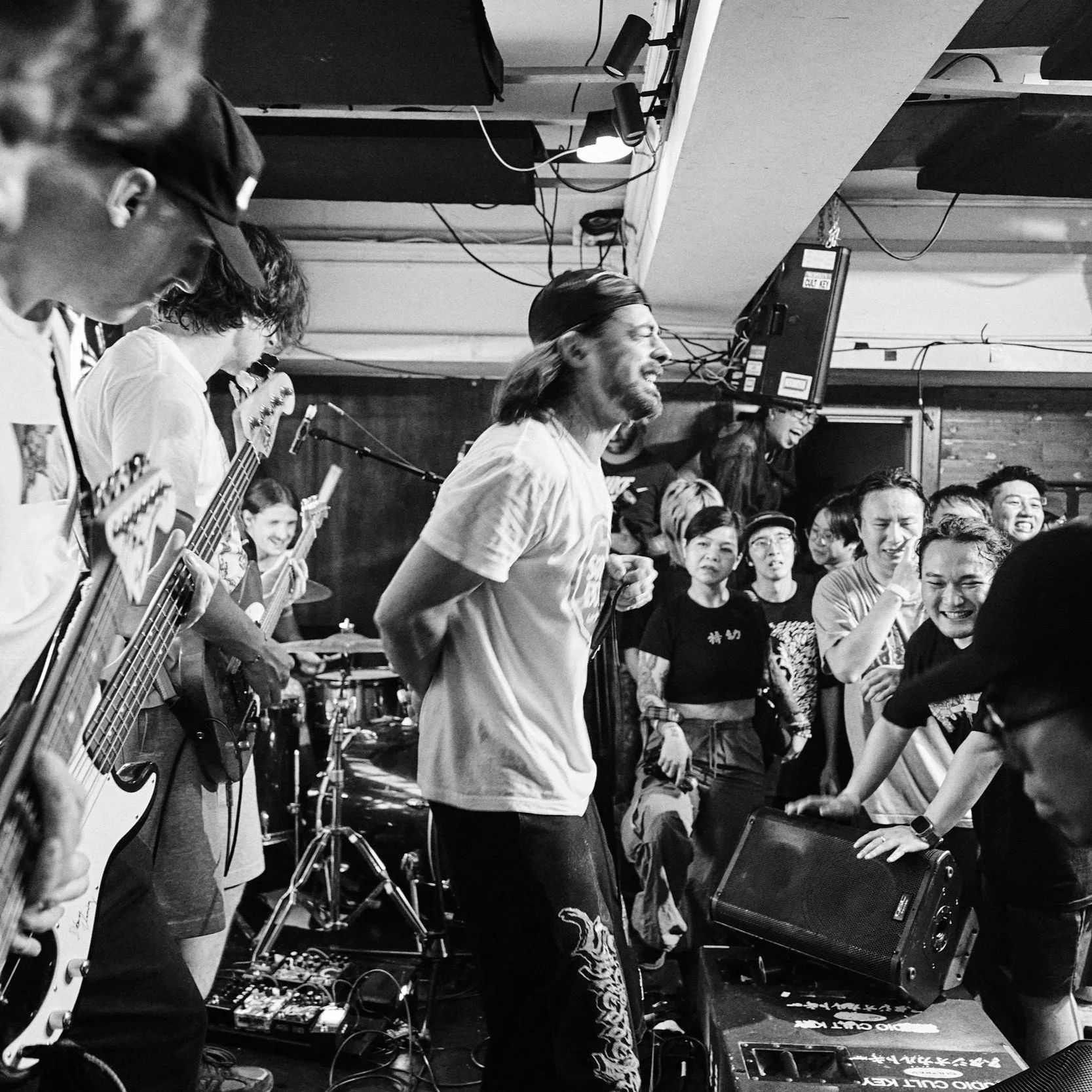 Black and white photo of musicians performing on stage, including a guitarist, a bassist, and a keyboardist, with a crowd of people watching and enjoying the concert.
