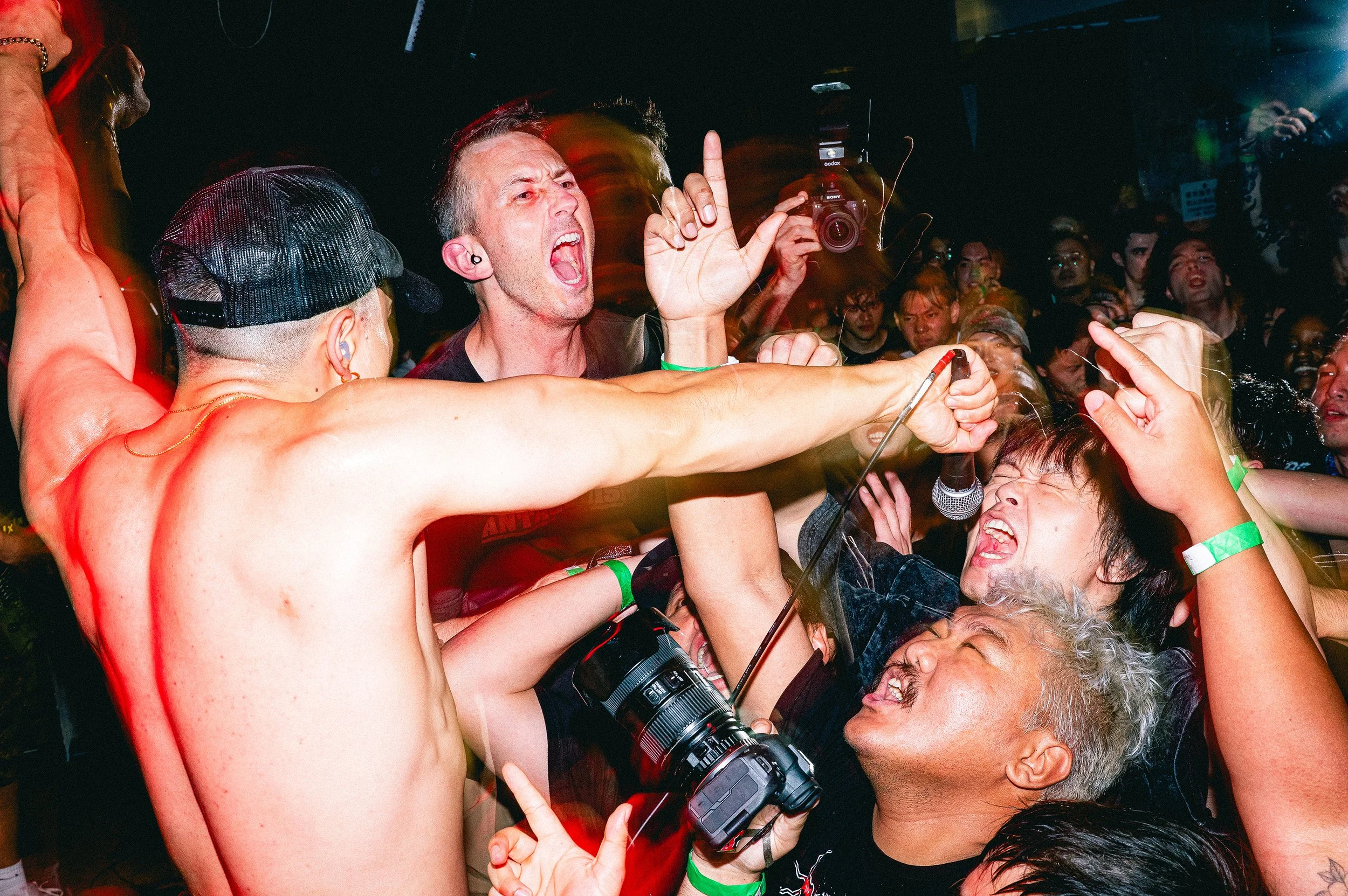 A group of people at a lively concert, some with their arms raised, passionately singing or shouting, with a man in the center making a loud vocal expression, surrounded by a diverse crowd amid dark lighting with red and green accents.