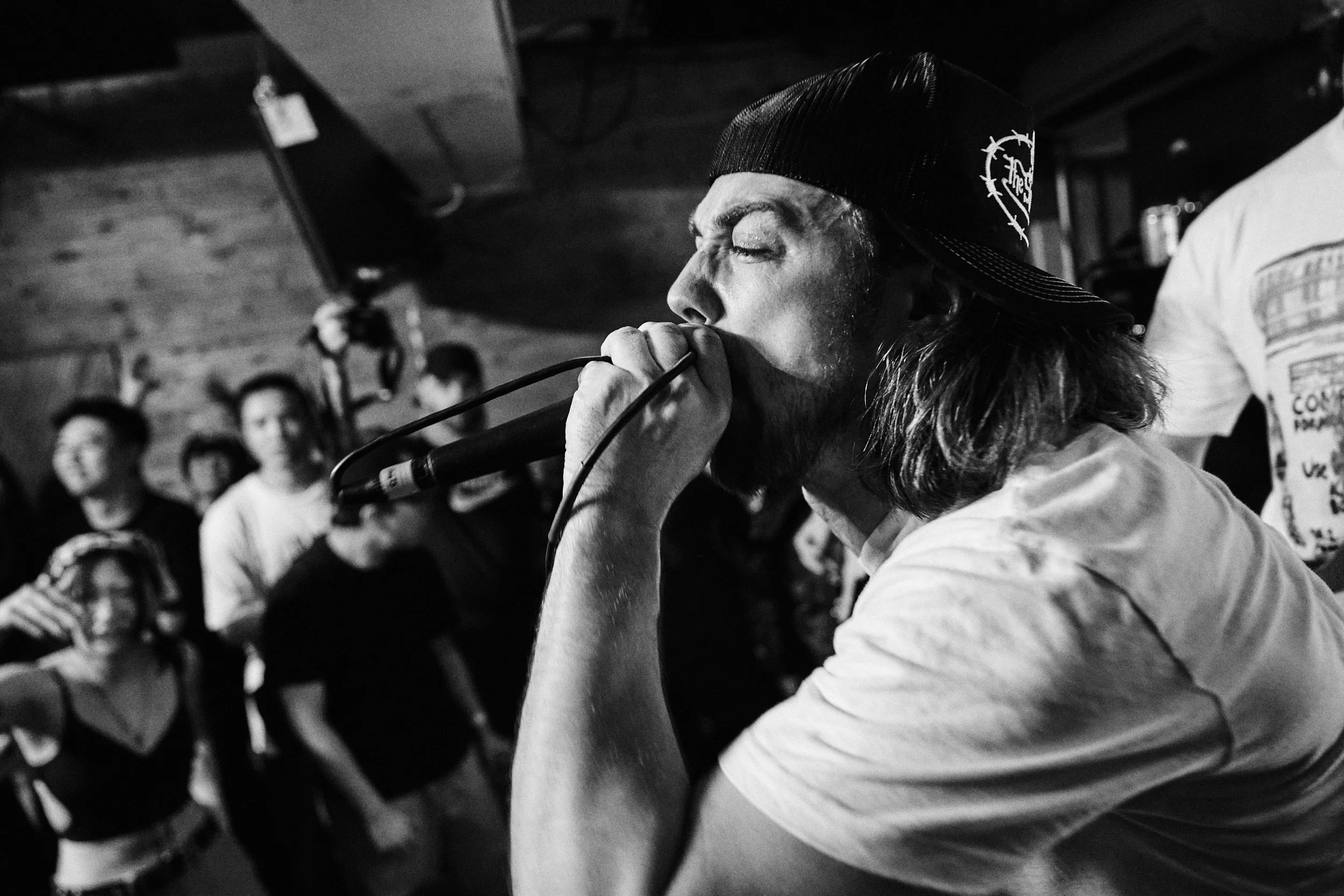 A person with long hair and a beanie hat singing into a microphone in a dimly lit room, with a blurred background of people.
