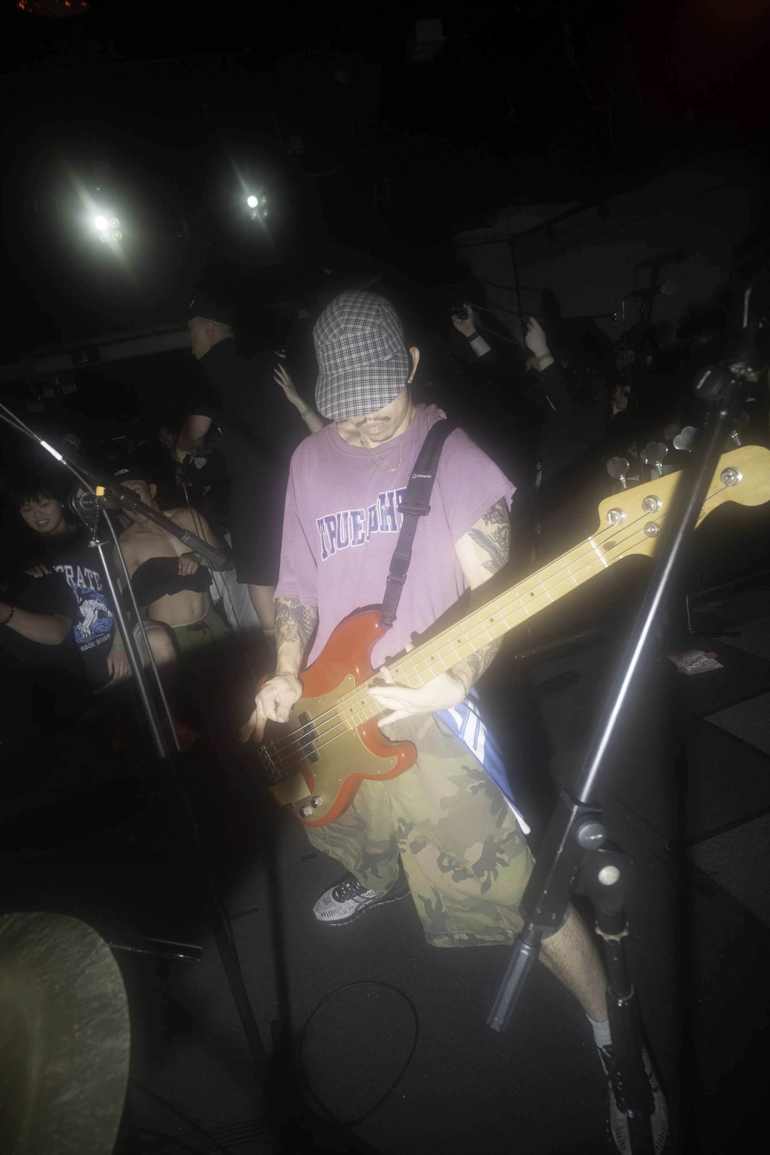 A man wearing a plaid cap, purple T-shirt, and camouflage shorts playing a bass guitar on stage with other people around him during a live music event.