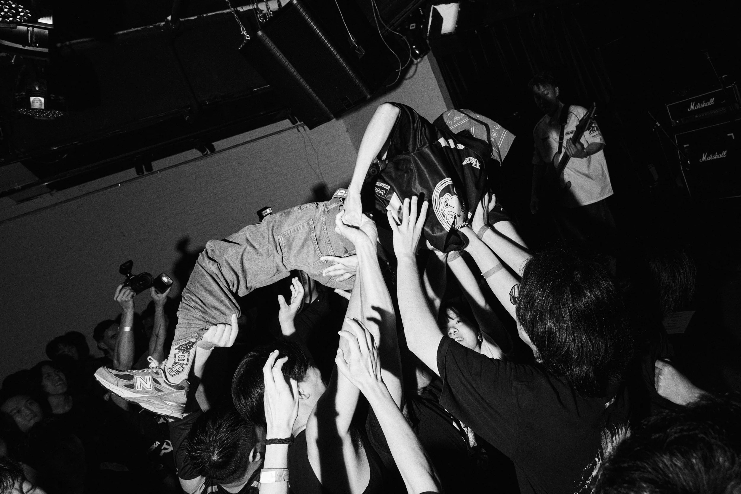 A black and white photo of a crowded concert or event where many hands are reaching out, with a person holding a microphone in the background.