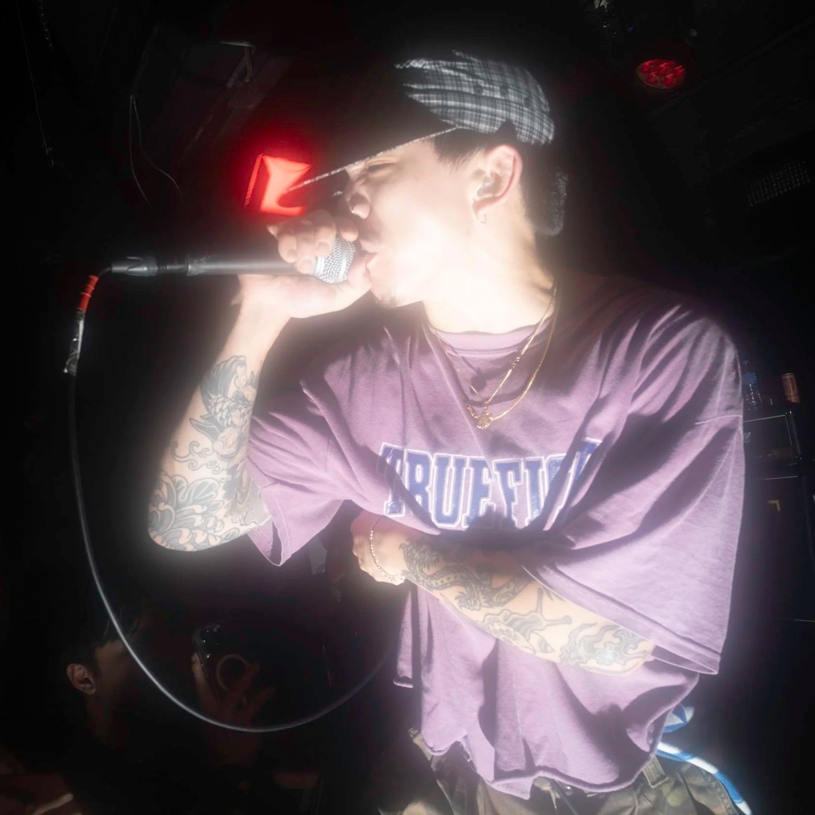A person singing into a microphone. The person has tattoos on their arms and is wearing a purple shirt with a graphic and layered necklaces. They have short hair under a checkered cap and are in a dimly lit setting.