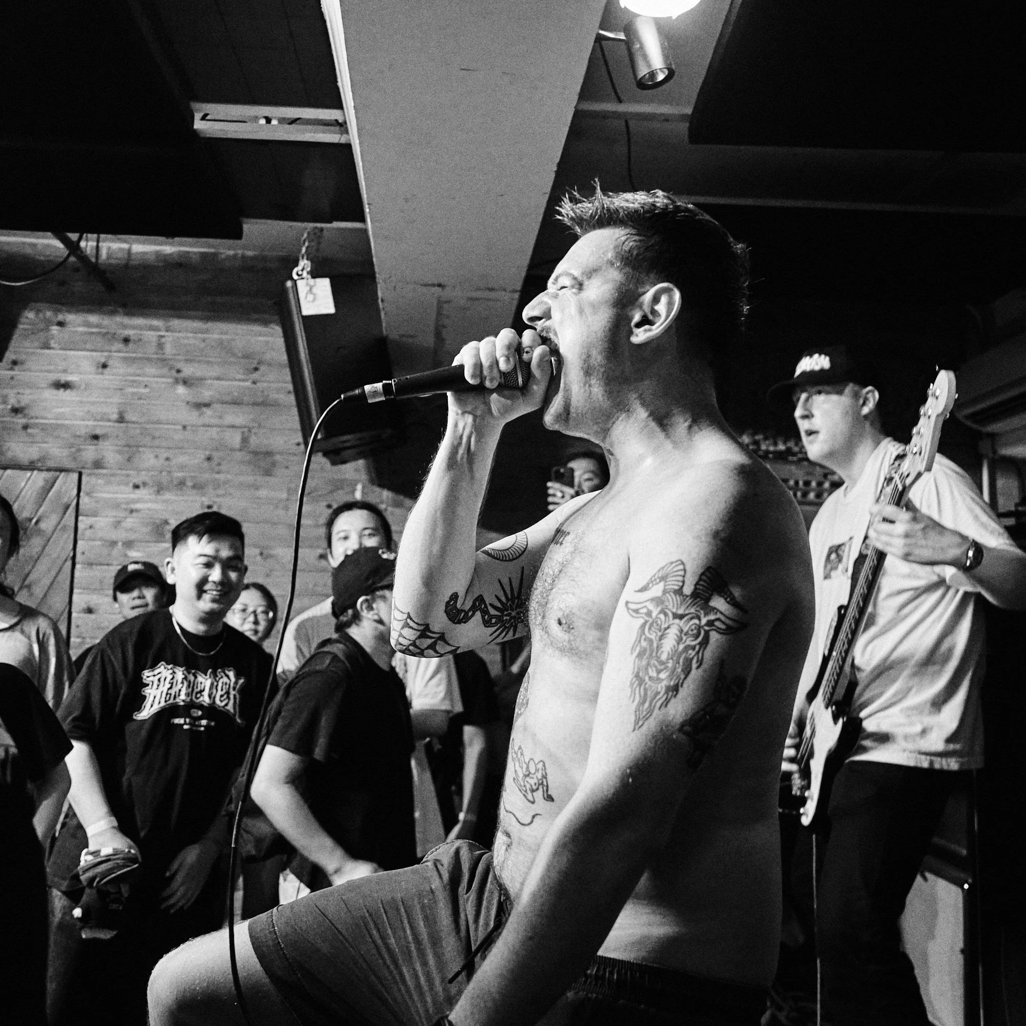 A black and white photo of a shirtless male singer performing with a microphone, tattoos visible on his chest and arms, wearing striped shorts and playing the guitar in a wooden, rustic venue with an audience watching.