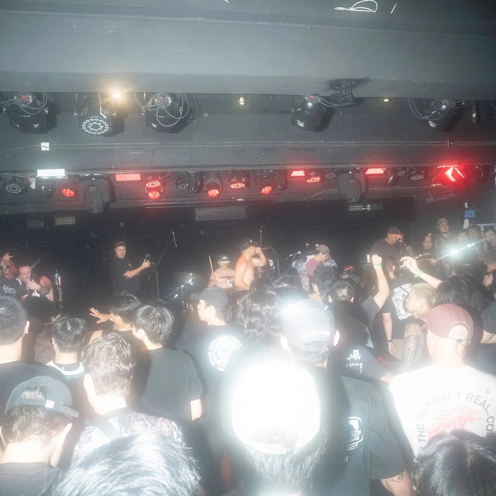 A live music concert with a band performing on stage, crowd of people watching, and stage lighting overhead.