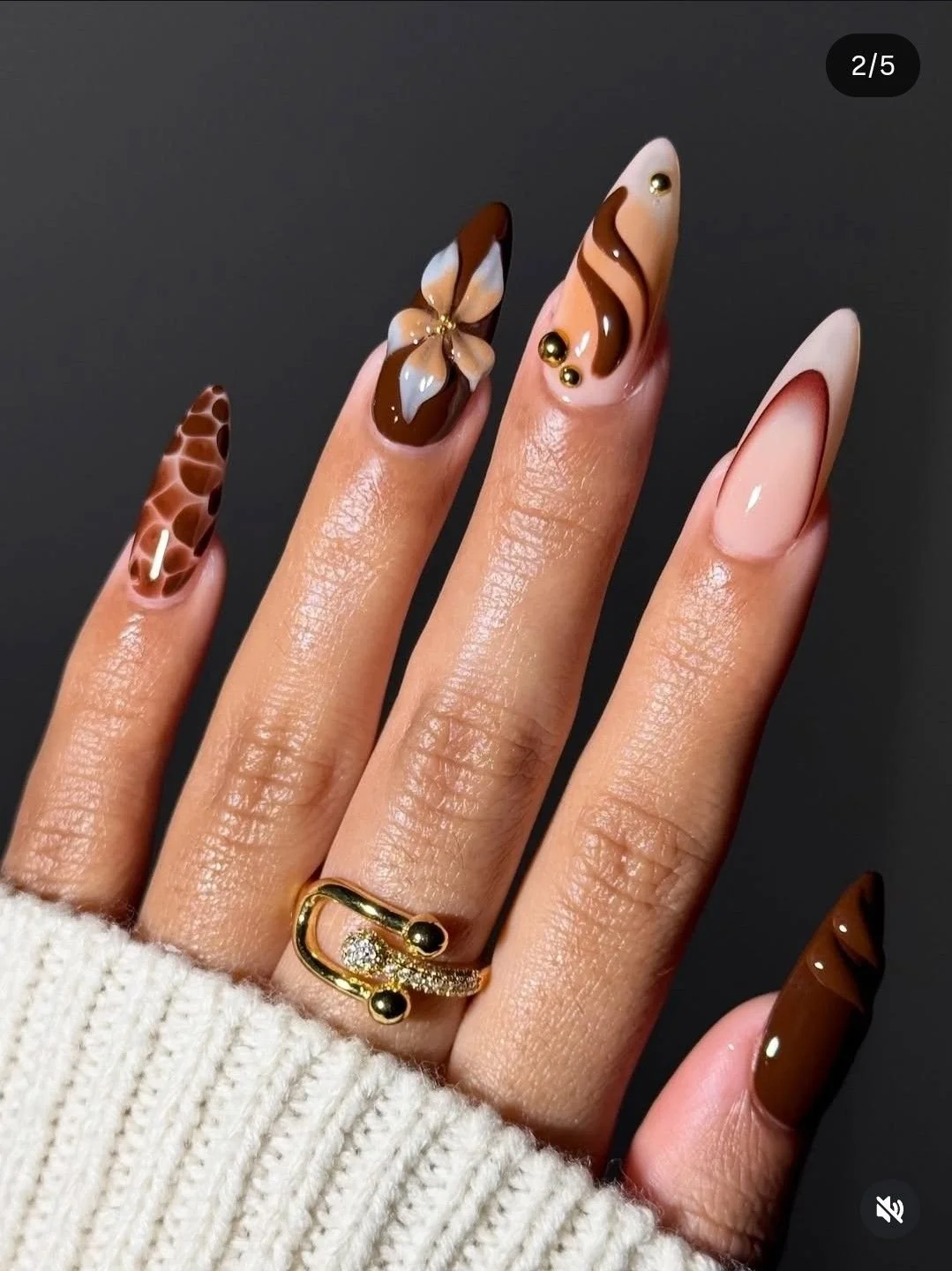 Nahaufnahmen einer Hand mit elaborierten, braunen und weißen Nageldesigns, goldenen Akzenten und einer goldenen Ring. Die Frau trägt eine cremefarbene Strickjacke.