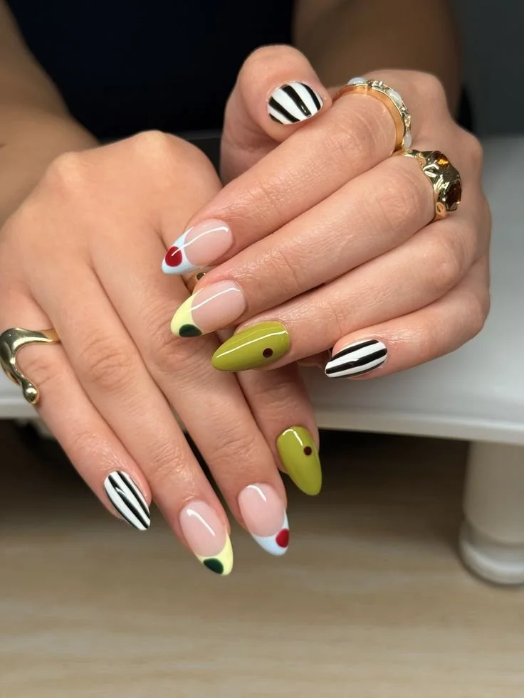 Nageldesign mit schwarzen und weißen Streifen, gelben, roten, schwarzen und grünen Akzenten. Hände mit Goldringen.