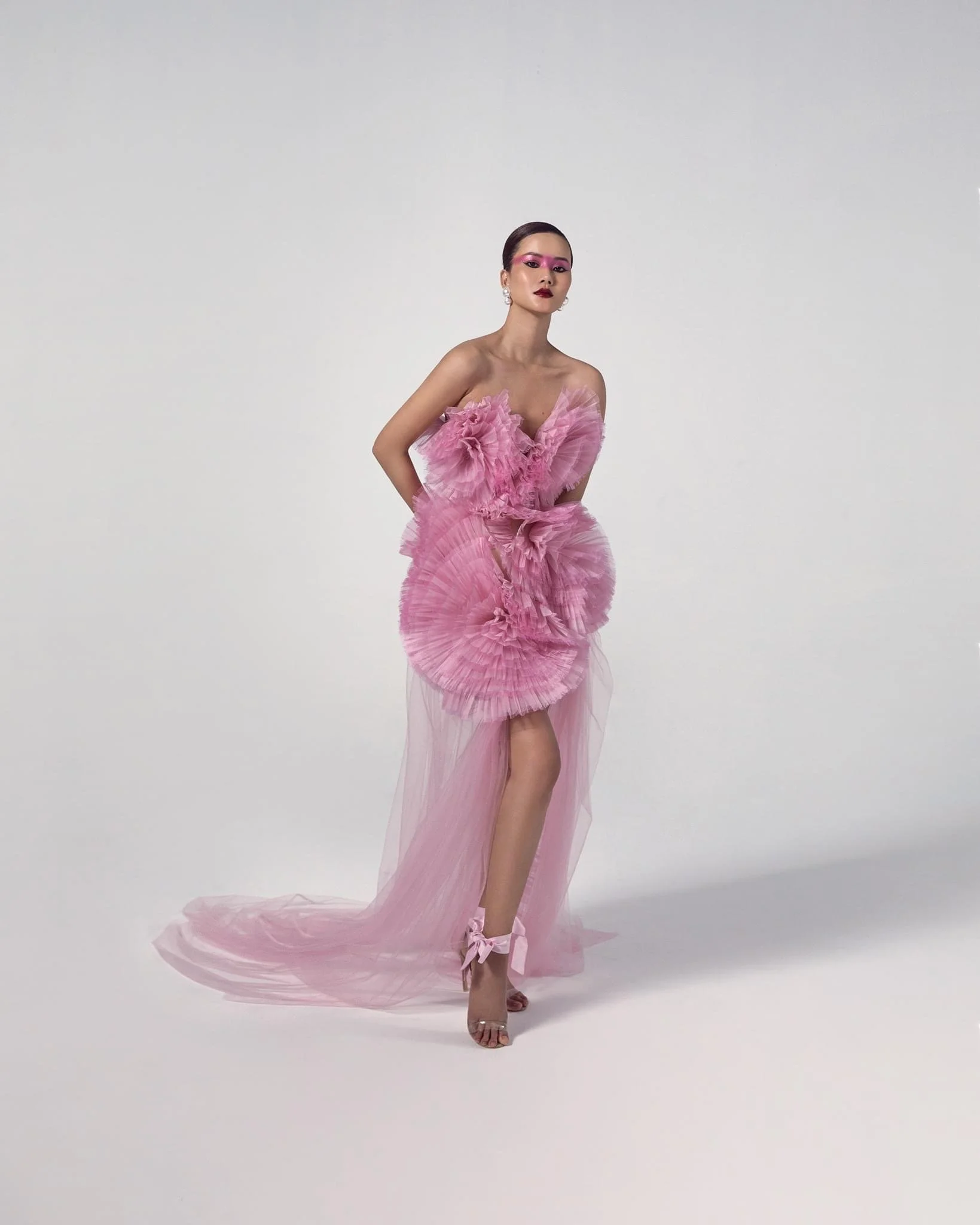 A woman in a pink, ruffled, sheer dress with a train, wearing high heels with bows, standing against a plain light-colored background.