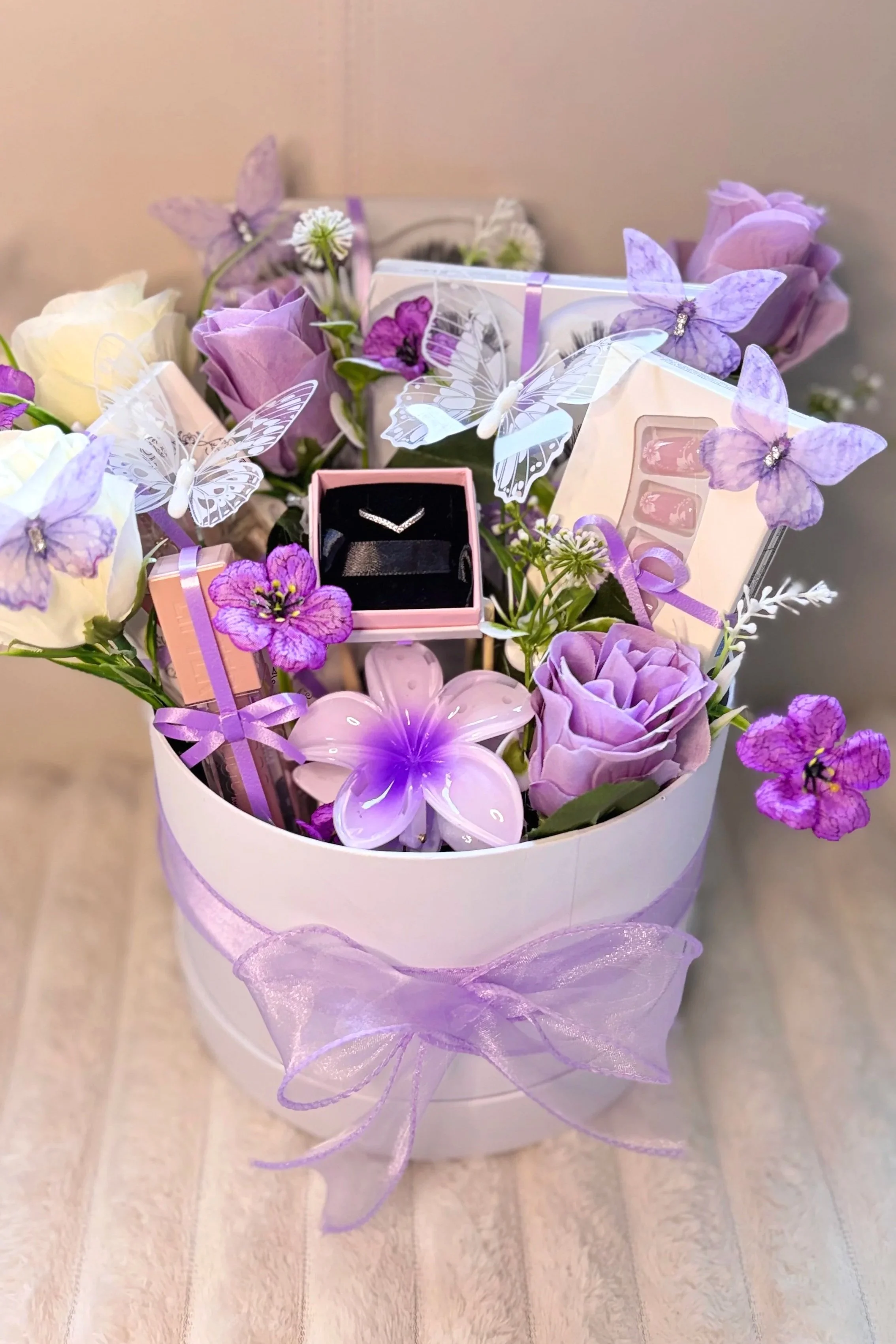 A white gift box with purple ribbon, filled with purple and white flowers, decorative butterflies, and jewelry inside a pink box. The box is placed on a light wooden surface.