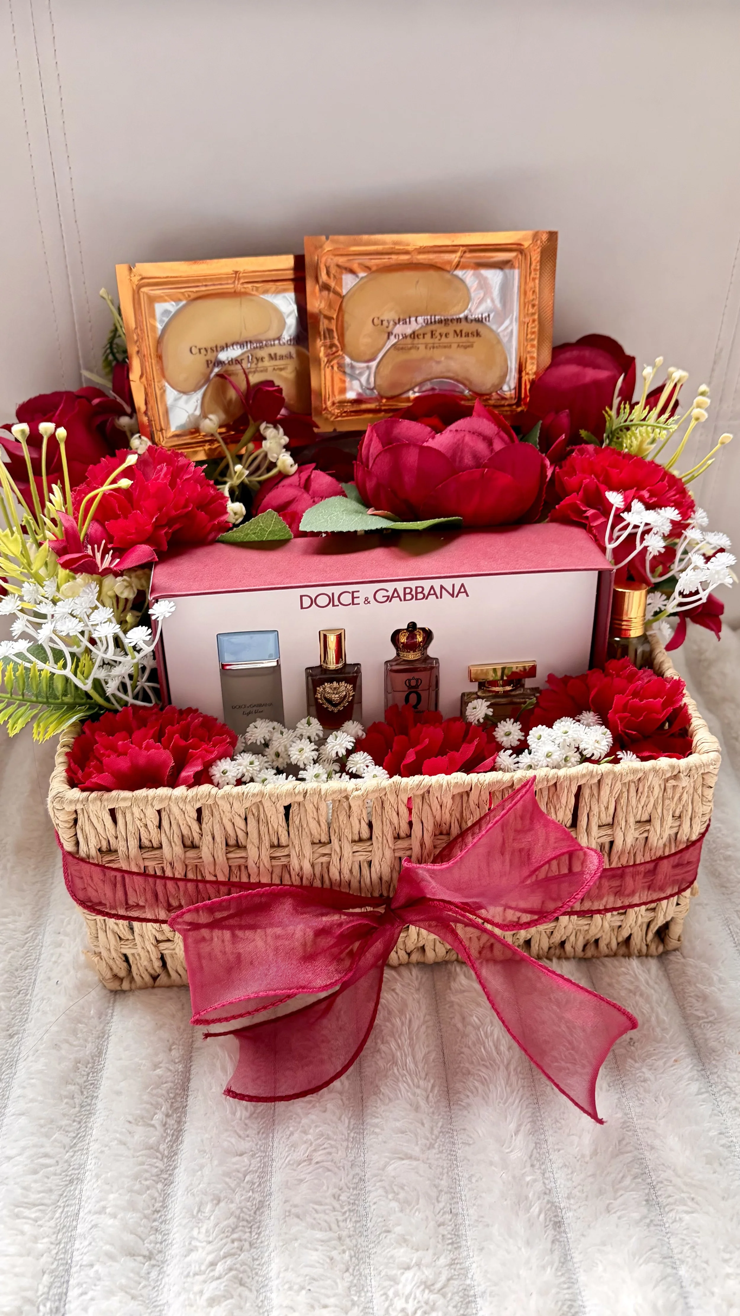A wicker gift basket decorated with a pink ribbon contains several bottles of perfume, surrounded by red, white, and pink artificial flowers, with two gold-coated face masks on top.