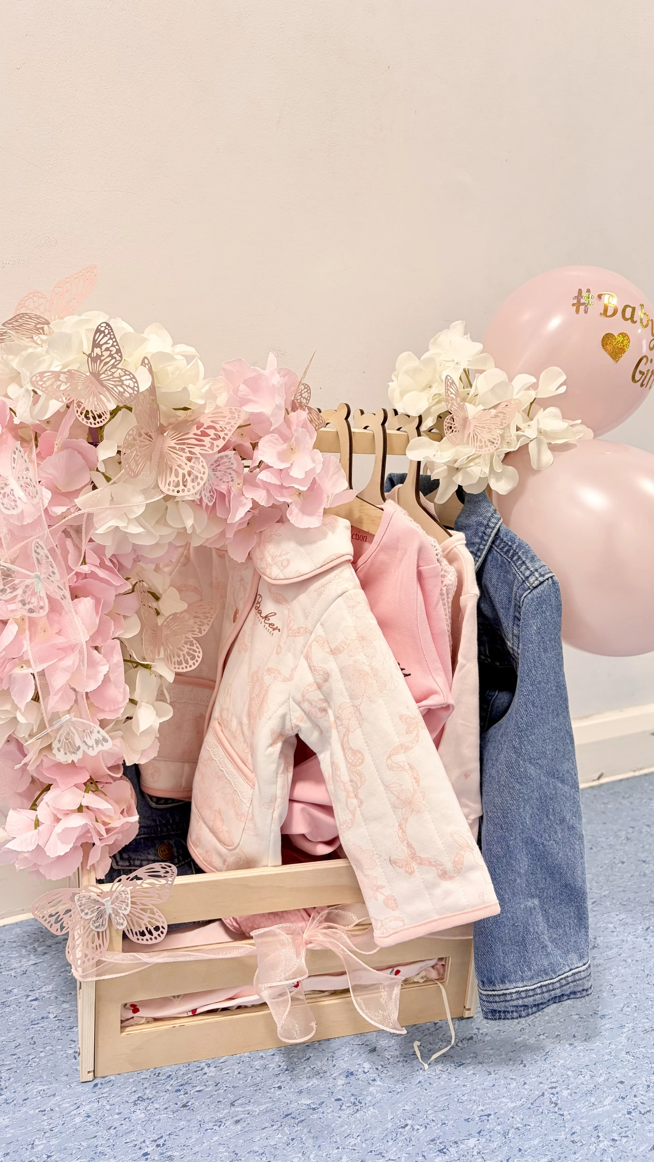 A wooden crate filled with pink and white baby clothes, surrounded by pink and white artificial flowers and butterflies, with pink balloons in the background decorated with the words '#BabyG' and a gold heart.