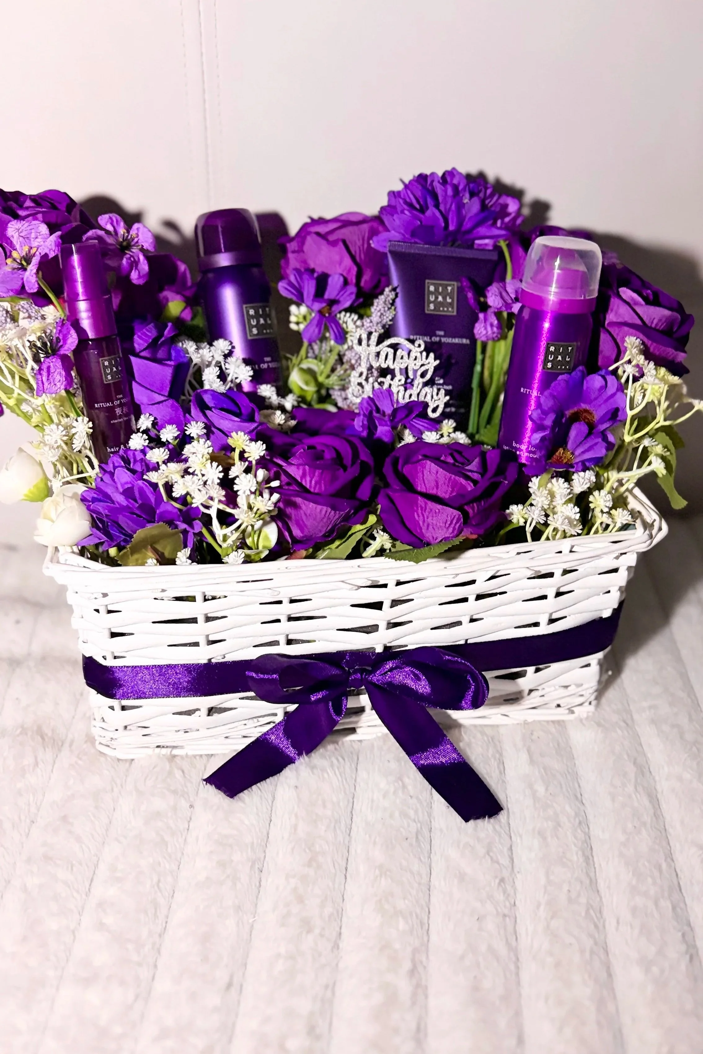 A white woven basket with purple ribbons, filled with purple and white flowers, containing purple bottles and a small black box with white writing.