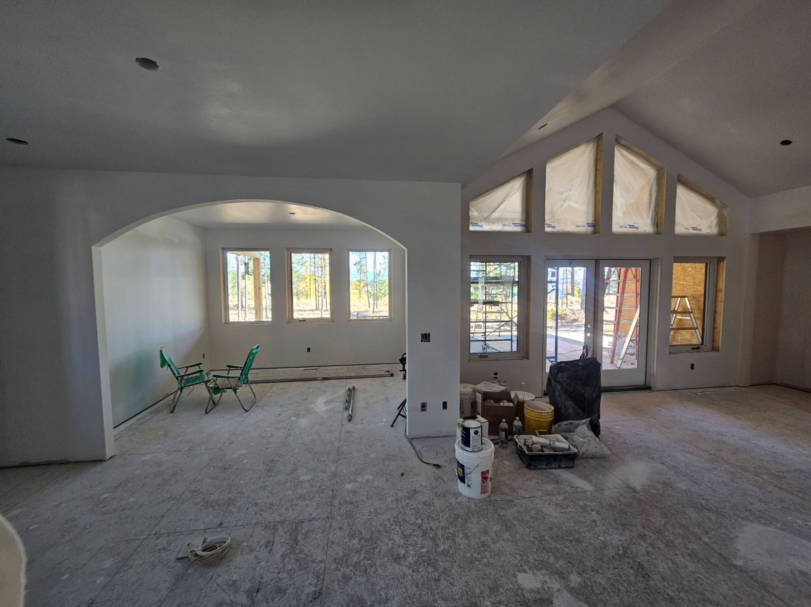Interior of a house under construction with unfinished floors, open wall spaces, and construction tools and supplies scattered around. Large windows and a double door leading outside are visible.