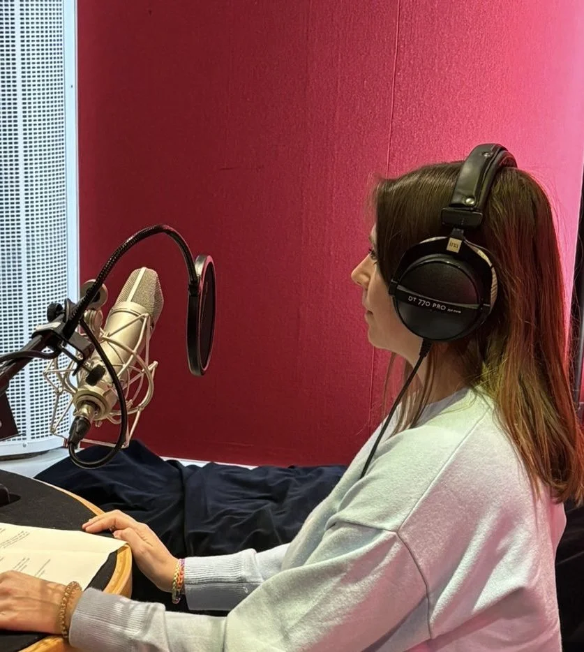 Une femme avec des écouteurs grand format assise devant un microphone dans un studio d'enregistrement, avec un fond rouge.