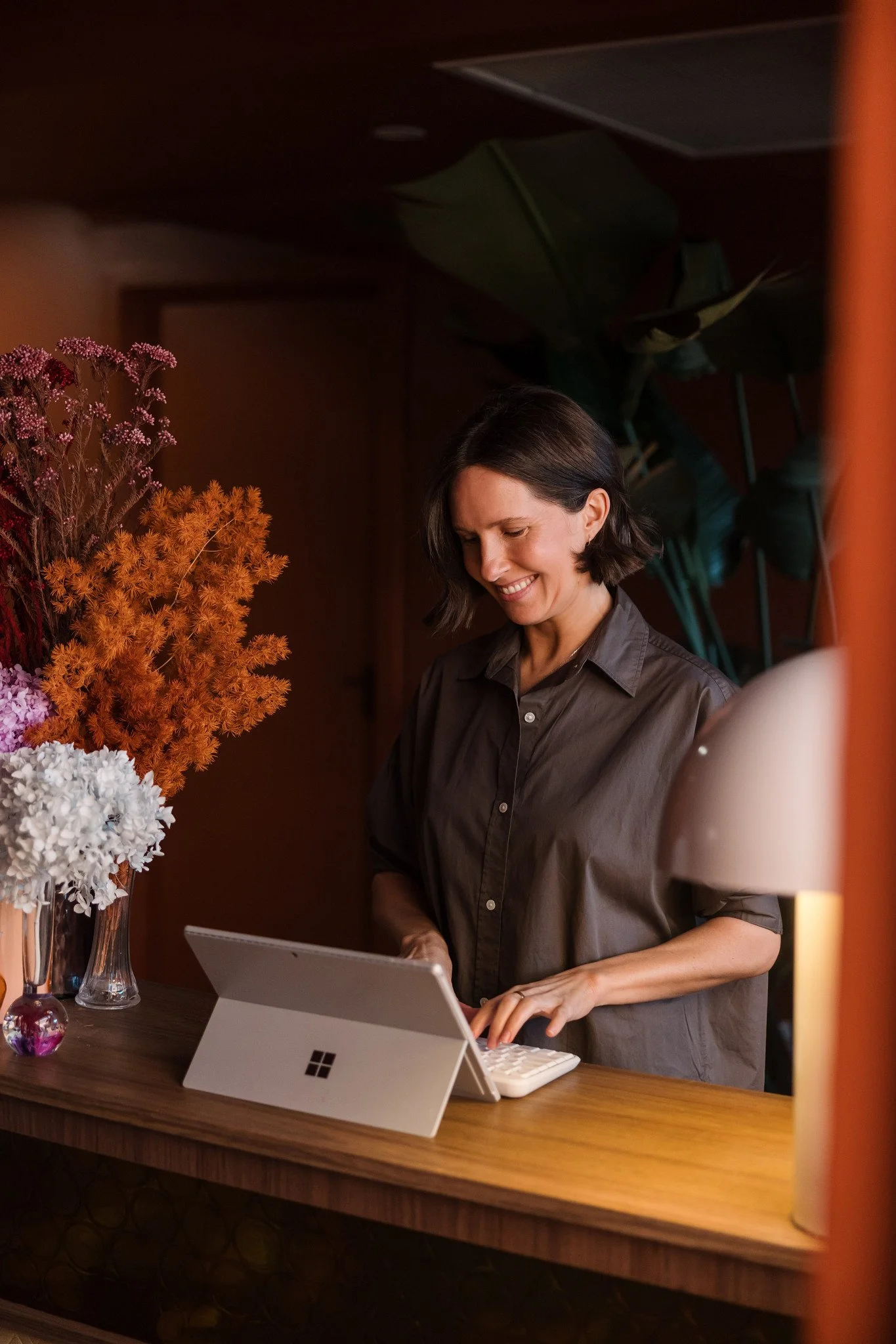 A woman in a gray shirt using a tablet or laptop at a wooden counter, with a vase of colorful flowers nearby, inside a warmly lit room.