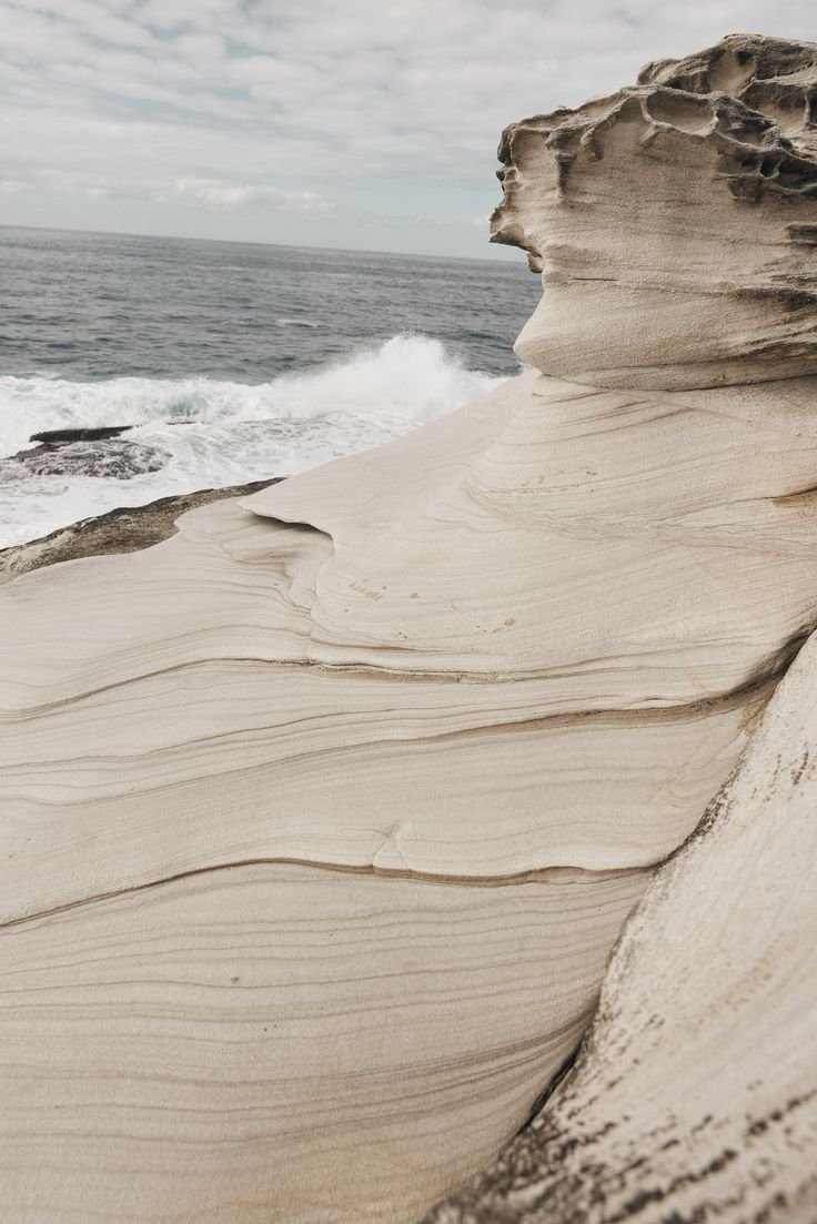 Close-up of layered, weathered sandstone formations along the ocean shoreline with waves crashing in the background under cloudy skies.