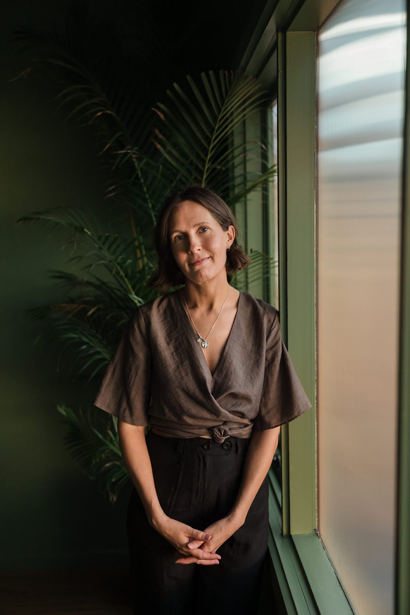 A woman in brown top and black pants standing by a window with green plants behind her.