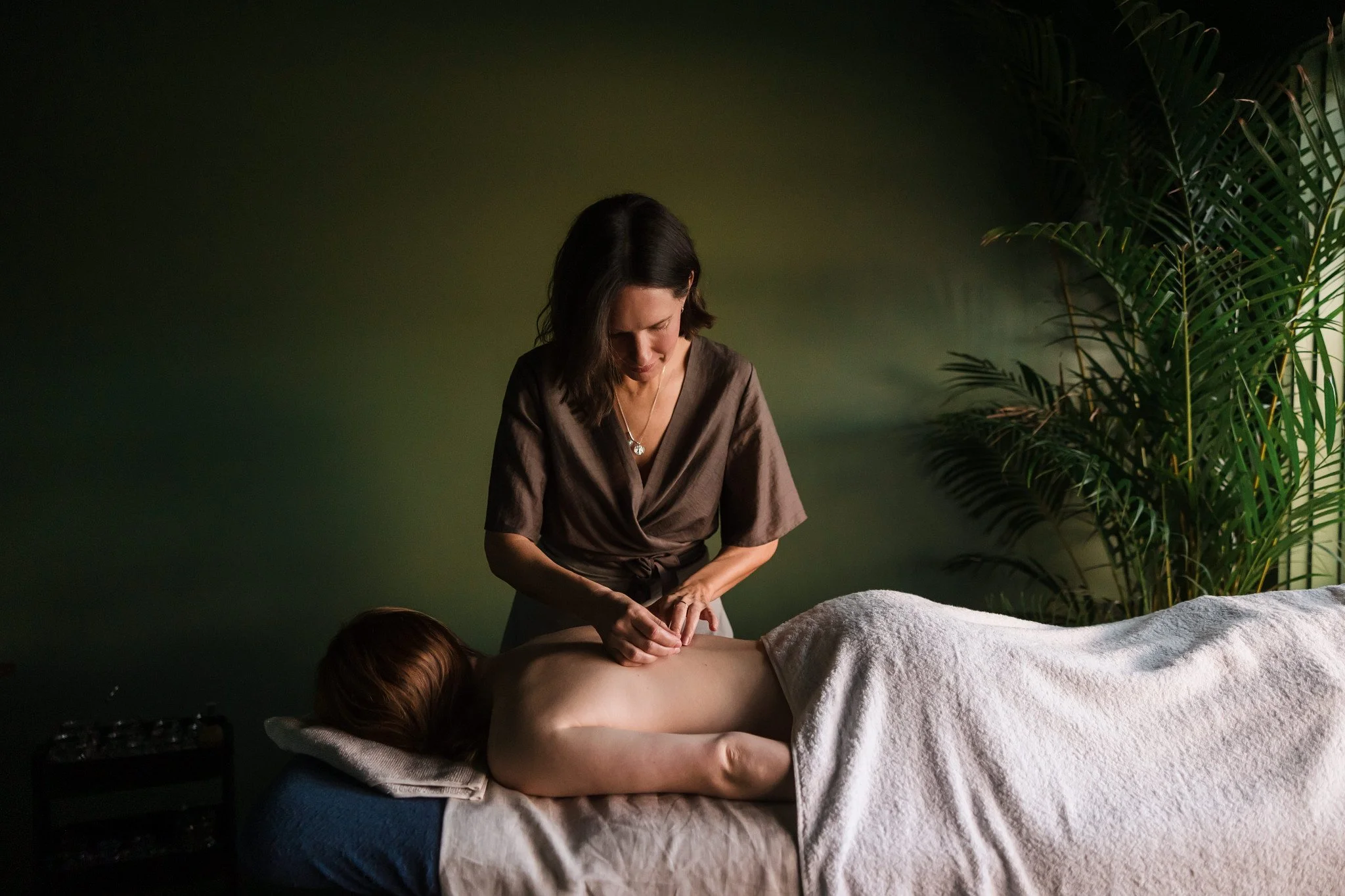 A woman giving a massage to a person lying face down on a massage table in a room with green walls and a large potted plant.