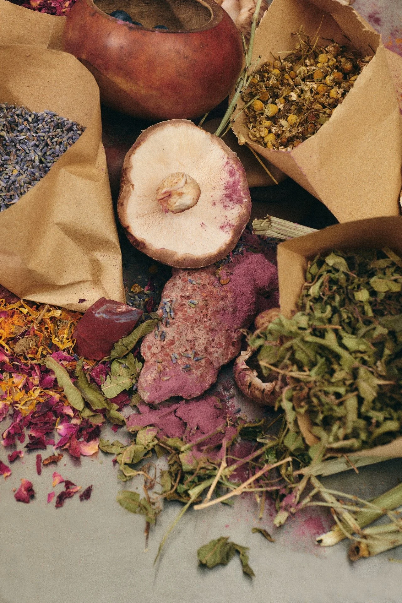 A variety of dried herbs, flowers, and natural ingredients displayed in paper bags and scattered on a surface.