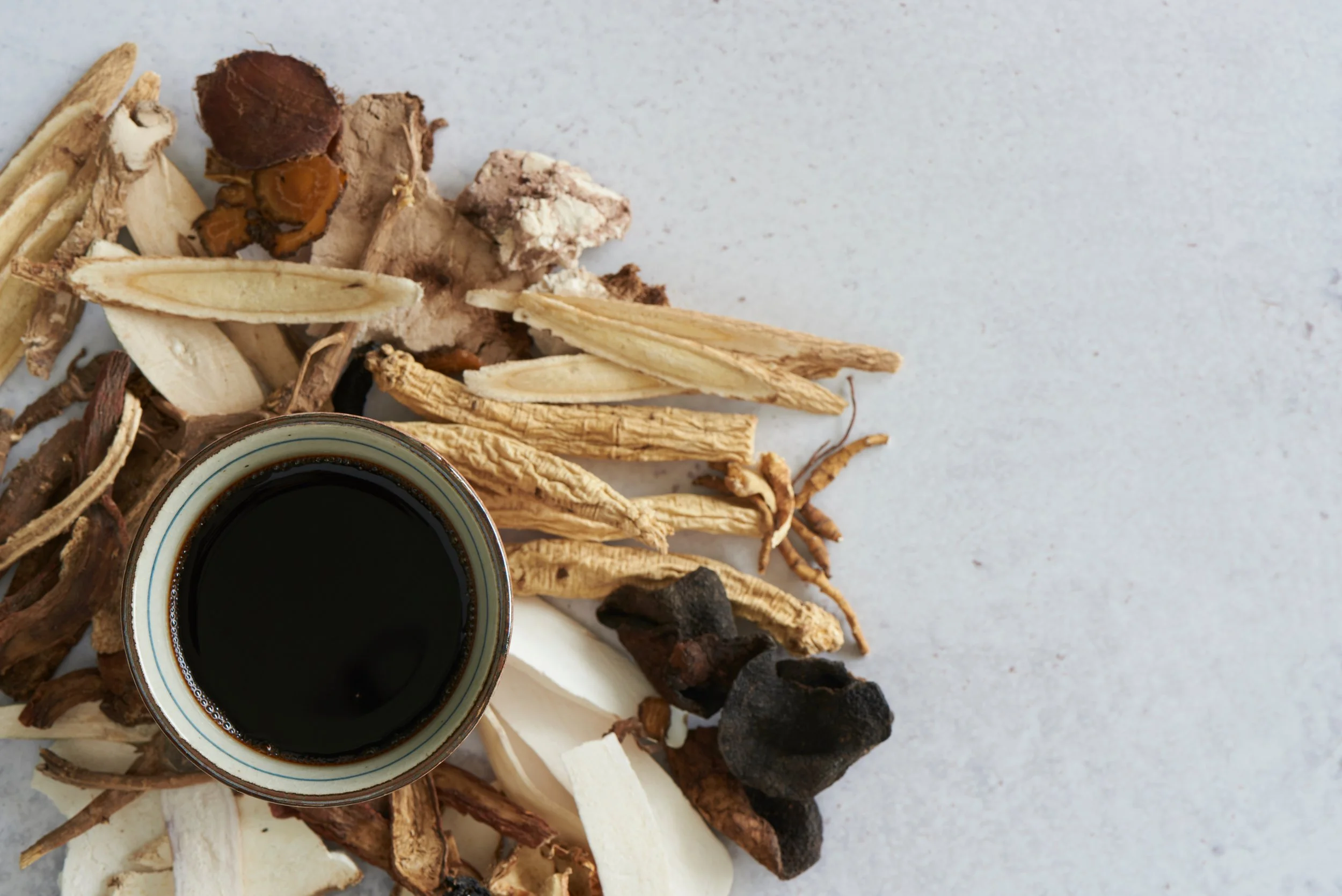 Cup of black Chinese herbal medicine, surrounded by assorted dried roots, herbs, and ginseng slices on a white surface.
