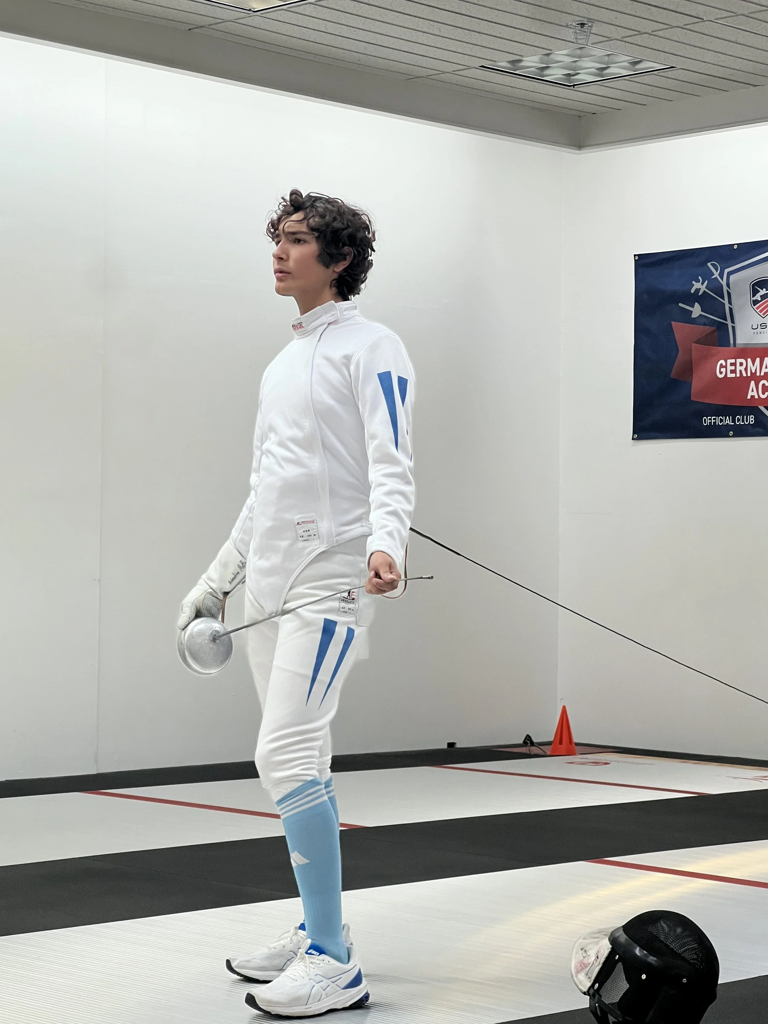 A young male fencer in white fencing gear, holding a fencing weapon, standing on a fencing strip in an indoor training facility, with fencing equipment and a banner in the background.