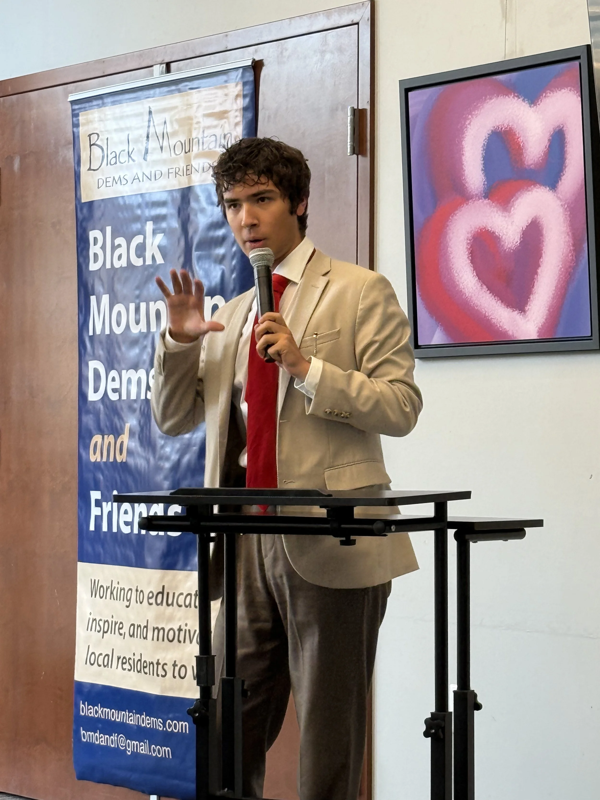 A man wearing a tan suit and red tie speaking into a microphone at a podium in front of a banner that says "Black Mountain Dems and Friends". There is a colorful artwork of hearts on the wall behind him.