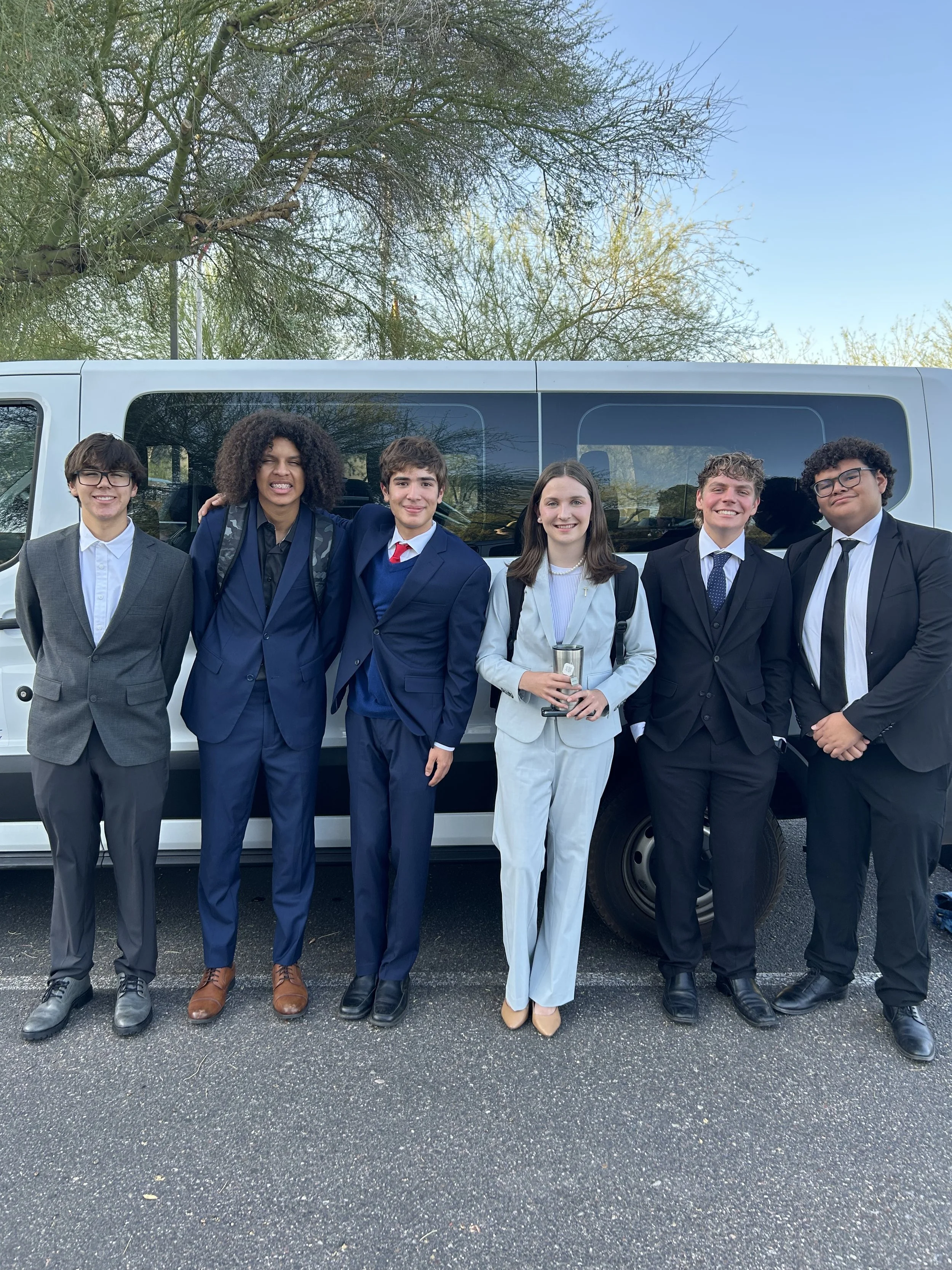 Group of six young people dressed in formal and semi-formal attire standing in front of a white van outdoors on a clear day.