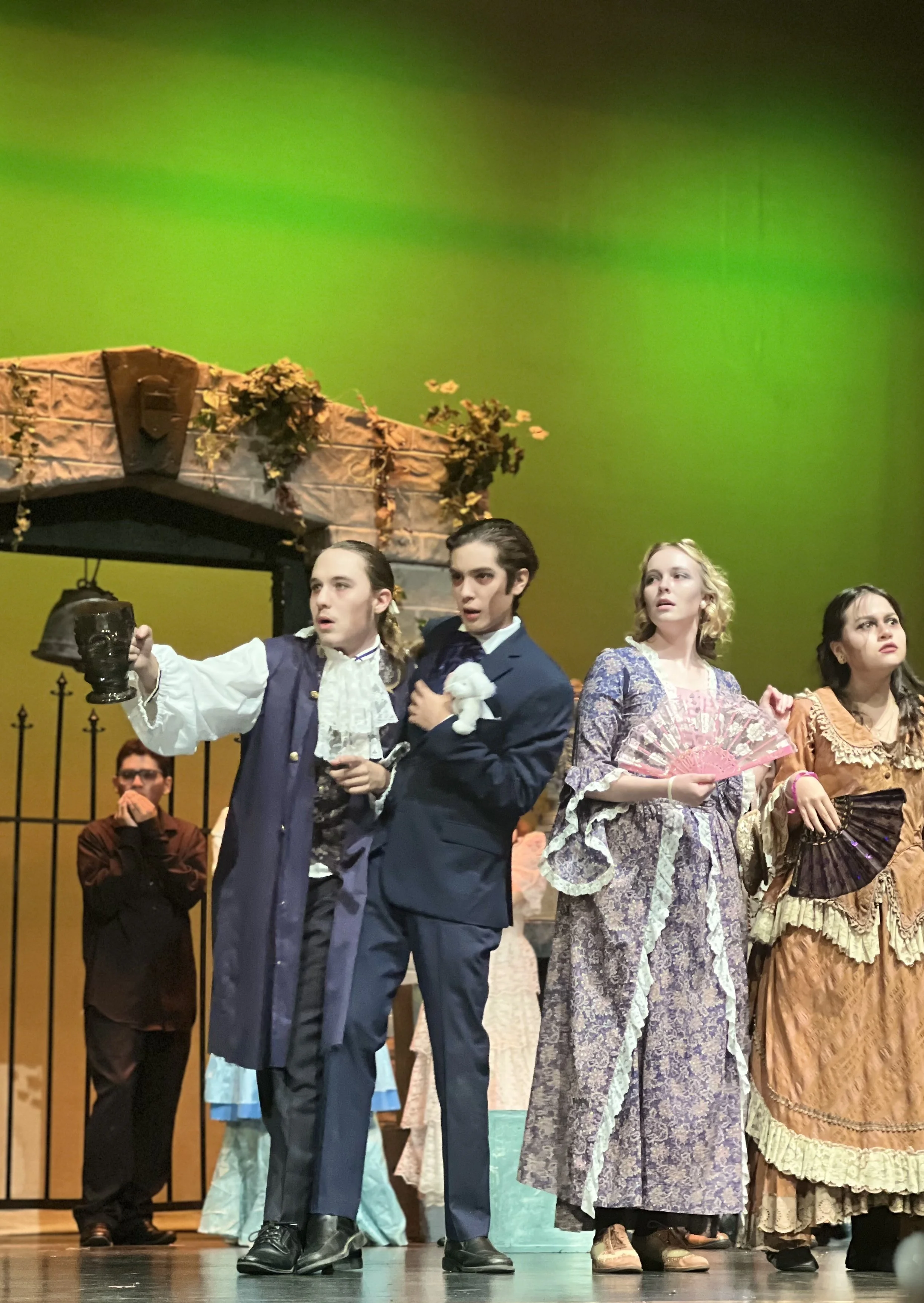 Actors in historical costumes performing on stage during a theatrical production, with a green backdrop and stage scenery.