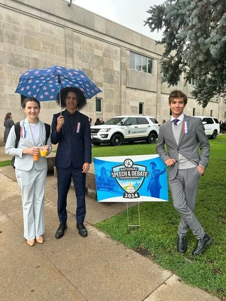 Three young people in formal attire standing outdoors near a sign for a speech and debate tournament in 2024. The person in the middle holds a blue umbrella with white stars. All three are smiling, and there are cars and a large stone building in the background.