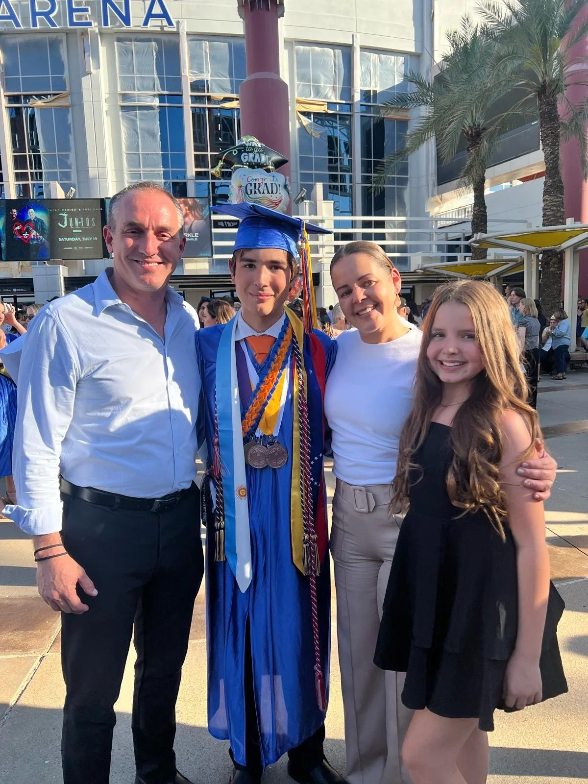 A family celebrating a graduate's accomplishment outdoors in front of a modern arena building with palm trees. The graduate wears a blue cap and gown with medals and cords, and the family poses happily around him.