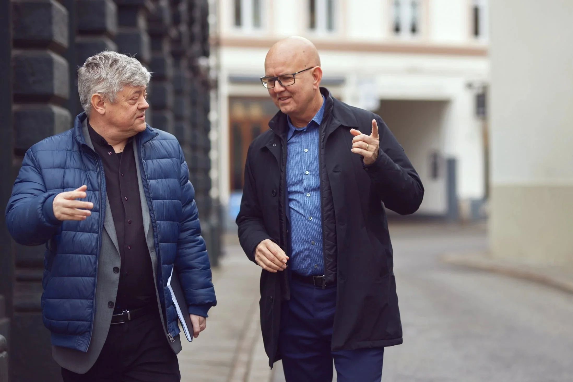 To mænd går snakker sammen på gaden, en med grå hår og blå jakke, den anden er bald og bærer briller og sort jakke.