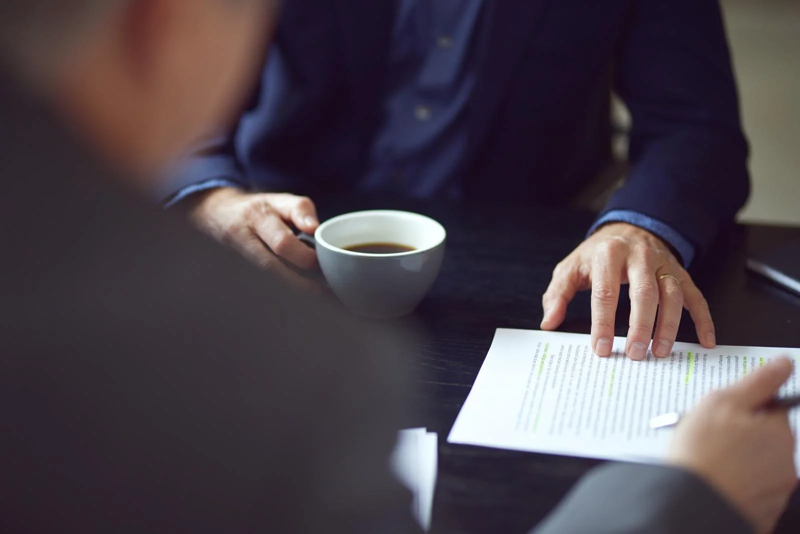 To personer sidder ved et bord, hvor en person holder en kop kaffe, og den anden peger på et dokument med markeringer.