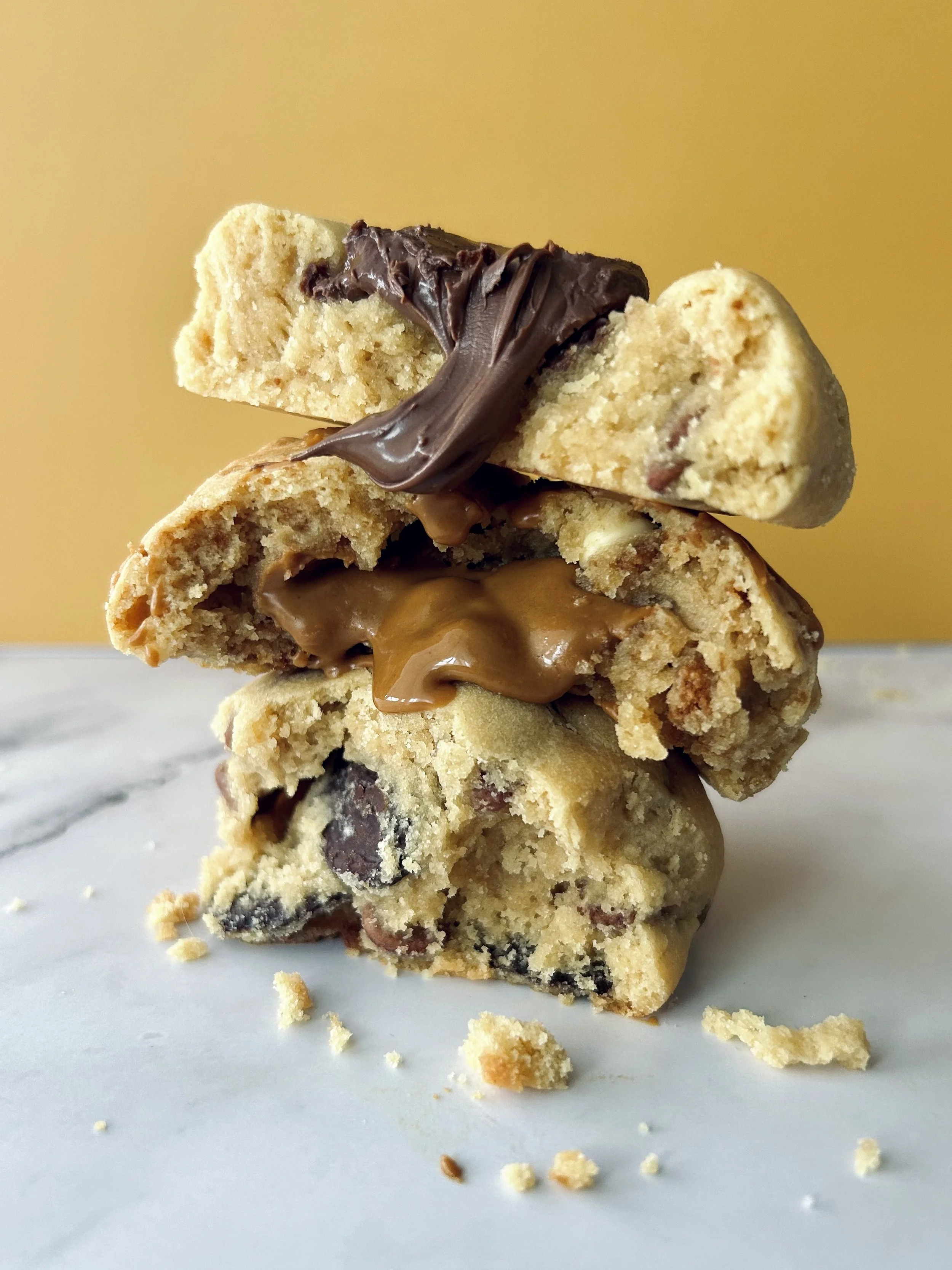Stack of three partially eaten cookies with chocolate and caramel, on a white surface with cookie crumbs, against a yellow background.
