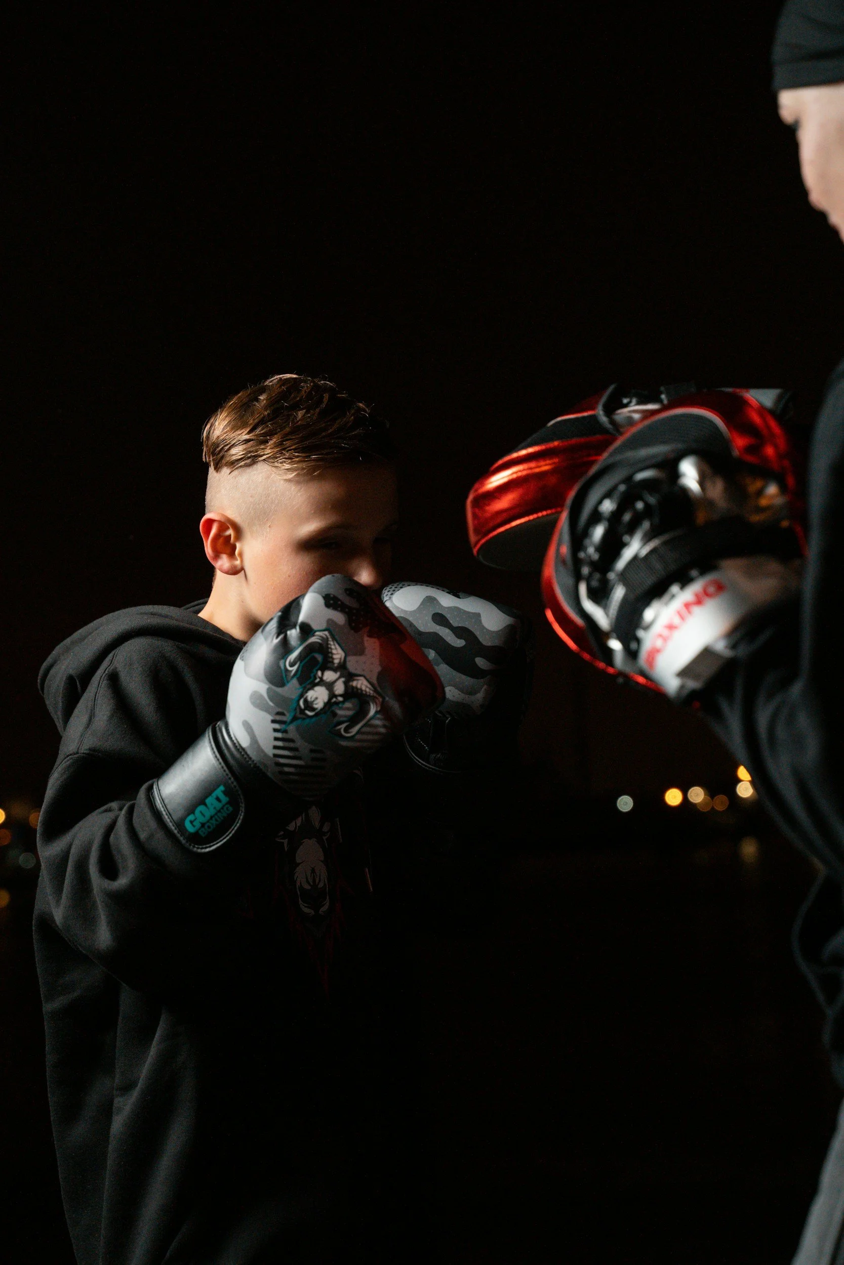 A young male boxer wearing boxing gloves, preparing to spar with another person, with a dark night background and some distant city lights.
