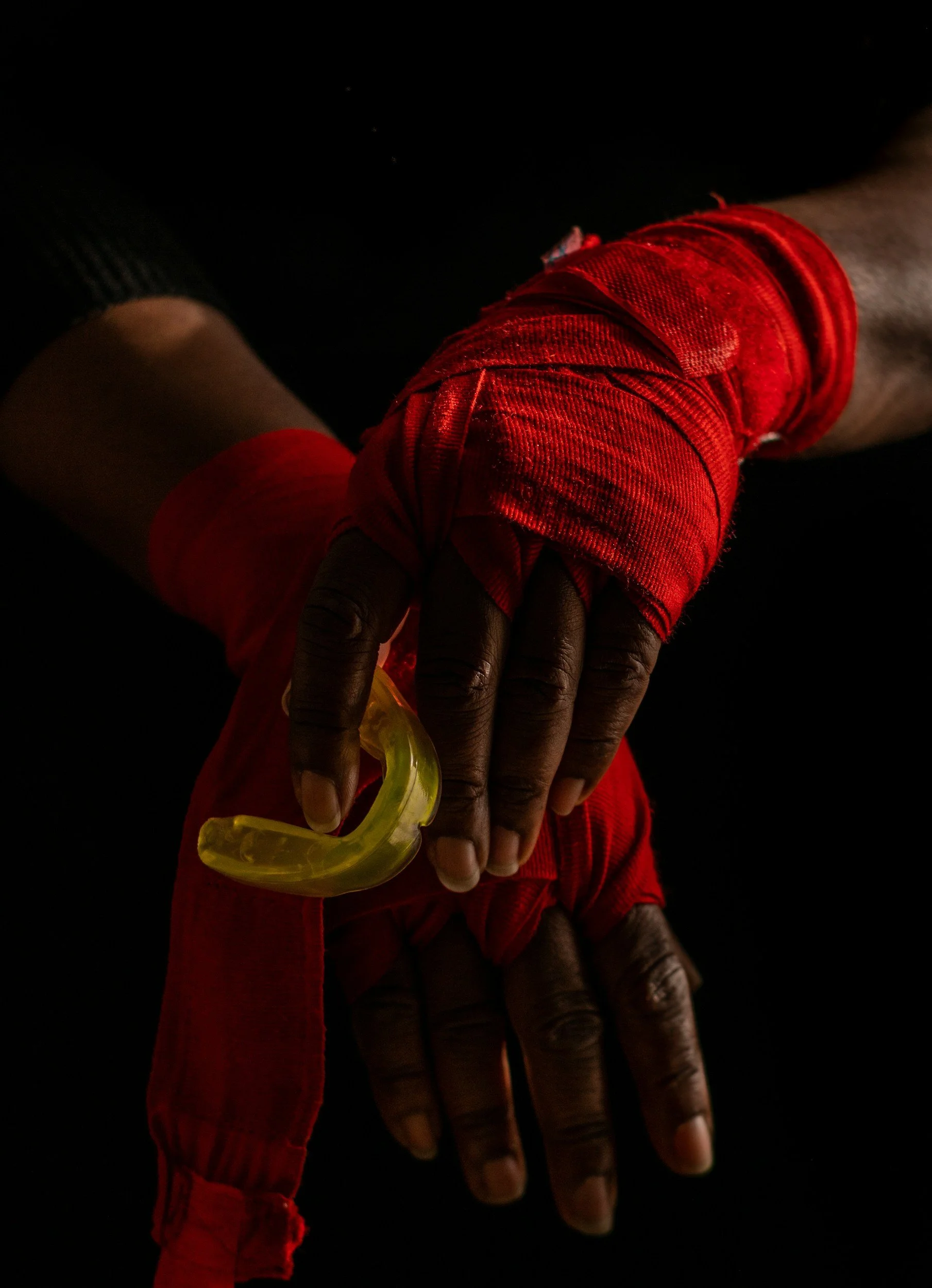 Close-up of dark-skinned hands, with one hand resting on top of the other, adorned with a yellow plastic ring and red cloth wristbands. The background is black.